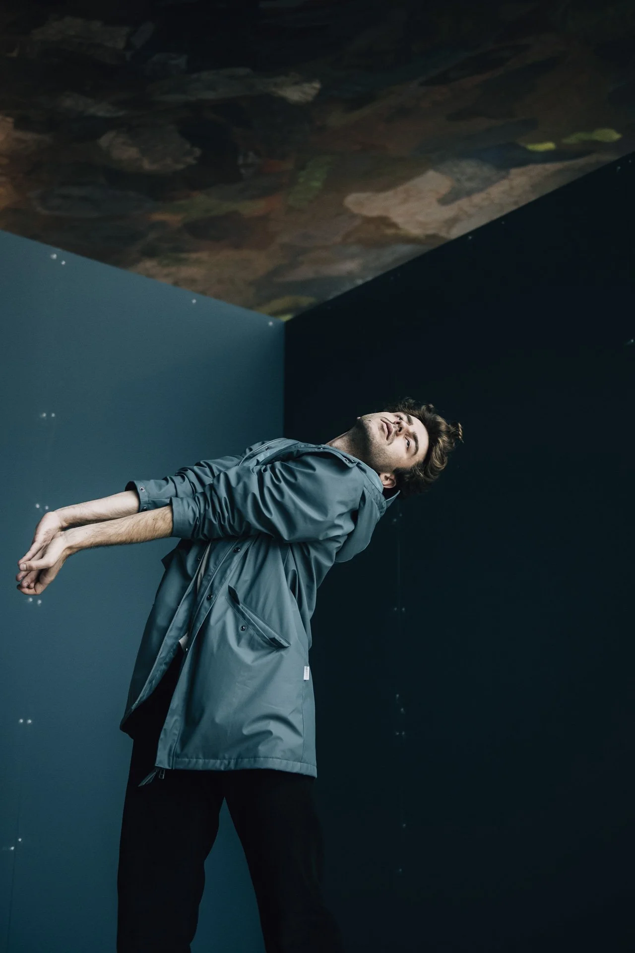 Ein junger Mann steht in einem dunklen Raum mit blauer Wand und Blick nach oben, während er eine graue Jacke trägt.
