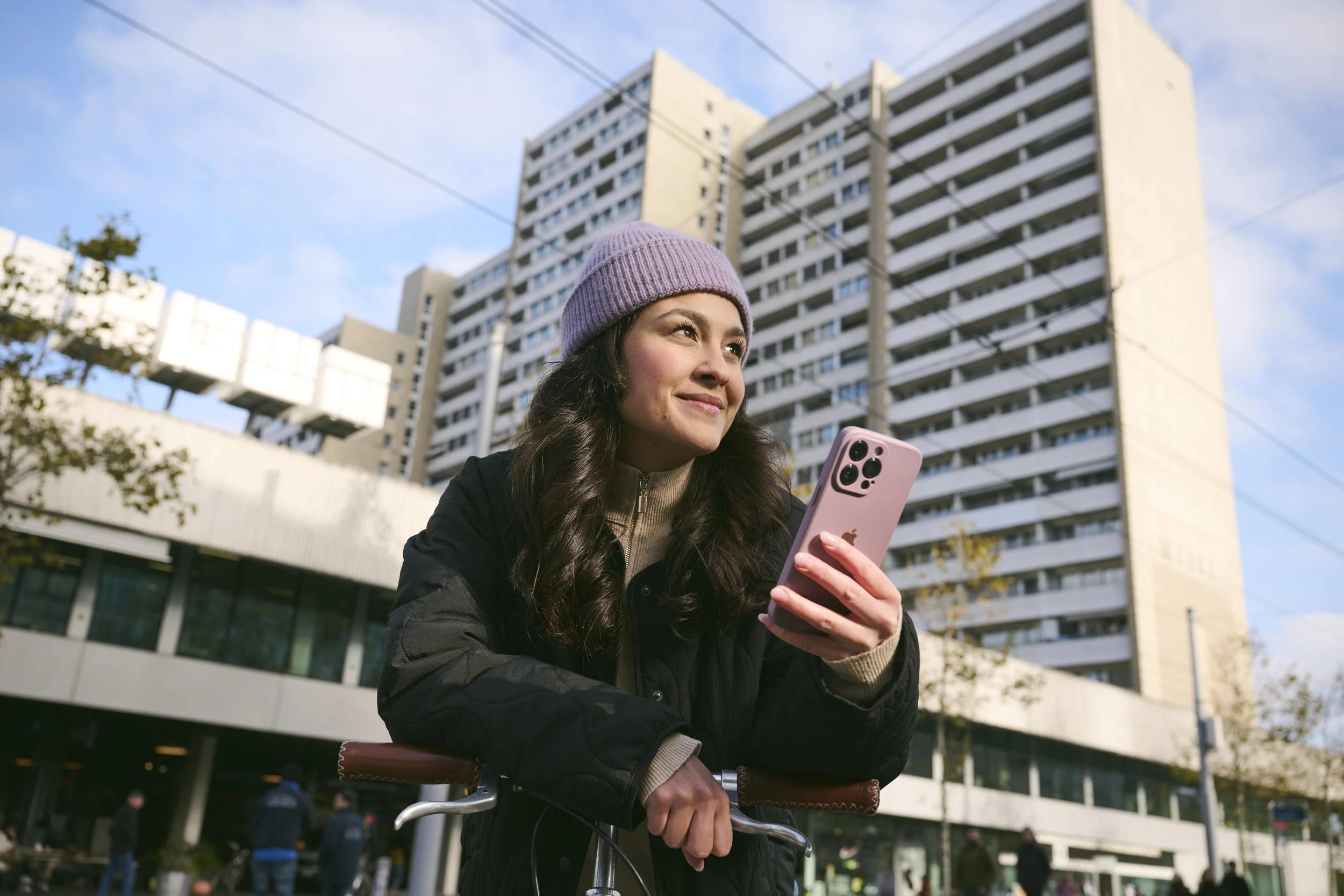 Eine Frau mit braunen Haaren, die eine rosa Mütze trägt, hält ein Smartphone und sitzt auf einem Fahrrad in einer modernen Stadtumgebung, im Hintergrund Hochhäuser und eine Gruppe von Menschen.
