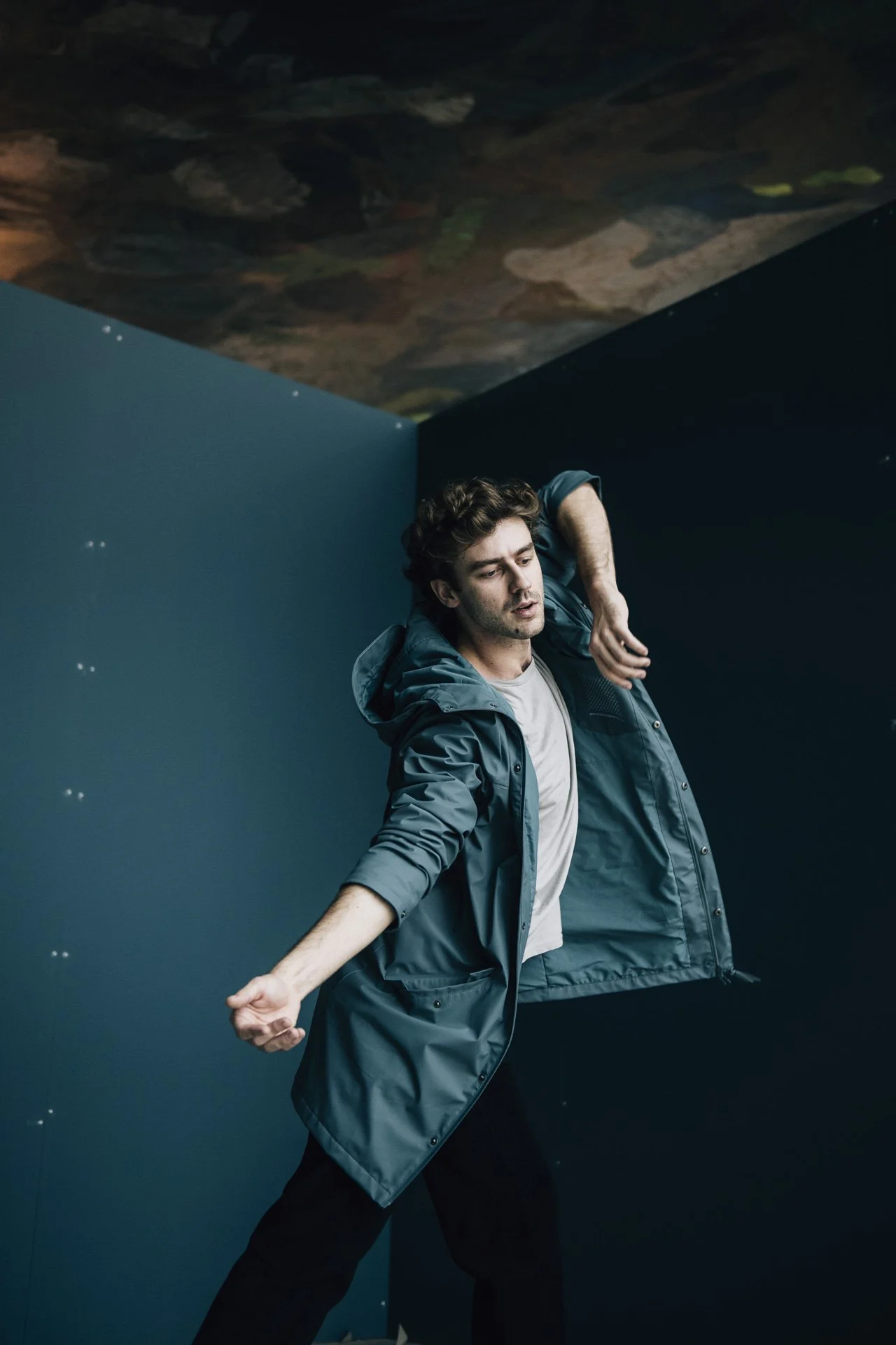 Ein junger Mann in einer dunklen Ecke, trägt eine graue Jacke, tanzt oder posiert mit ausgestreckter Armhaltung vor einer blauen Wand, im Hintergrund eine Deckeninstallation.