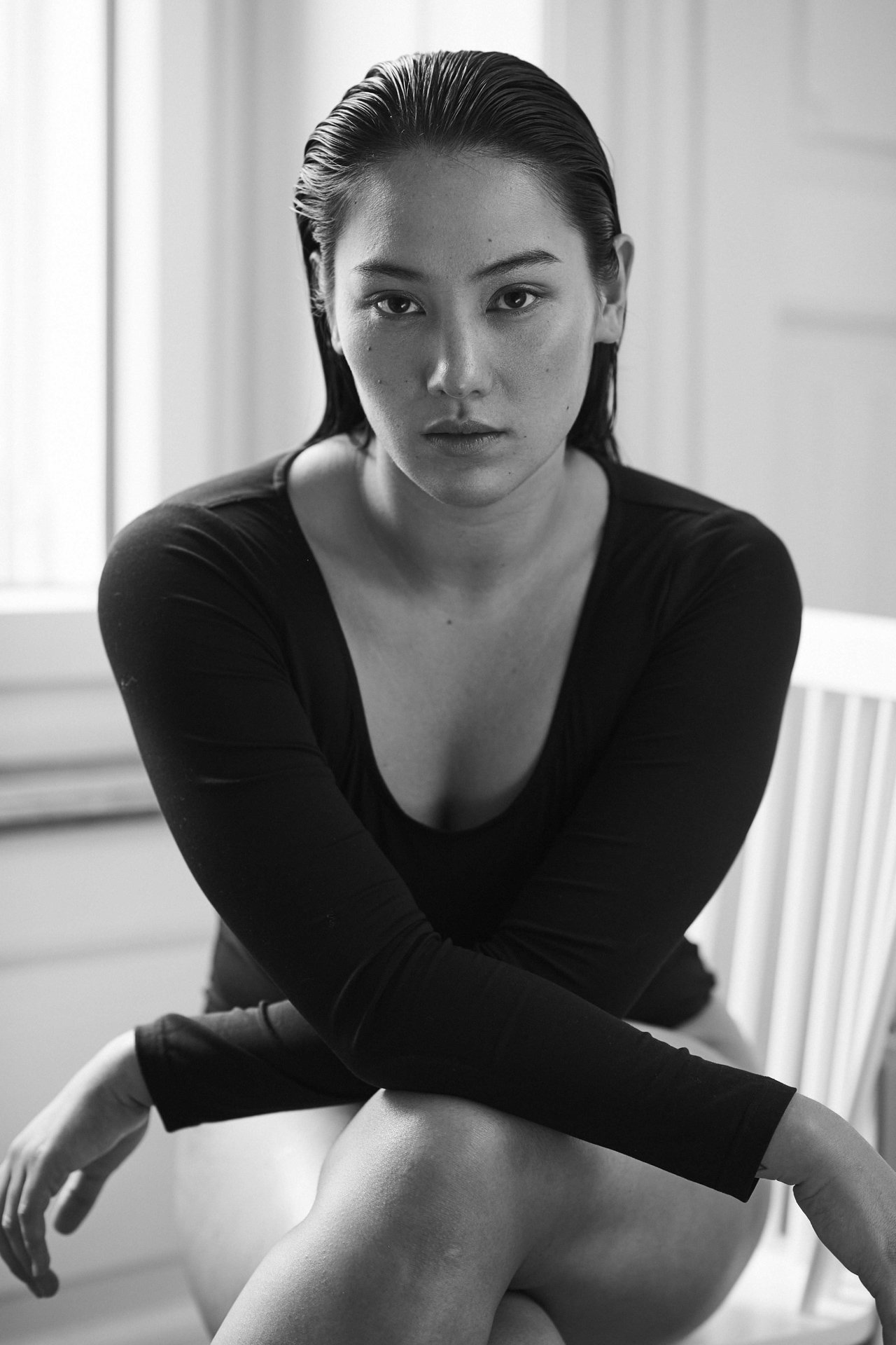 Schwarz-weiße Fotografie einer jungen Frau mit nassen, zurückgekämmten Haaren, sitzend auf einem Stuhl, mit Blick direkt in die Kamera. Sie trägt ein langärmliges schwarzes Oberteil und sitzt mit verschränkten Beinen vor einem neutralen Hintergrund.