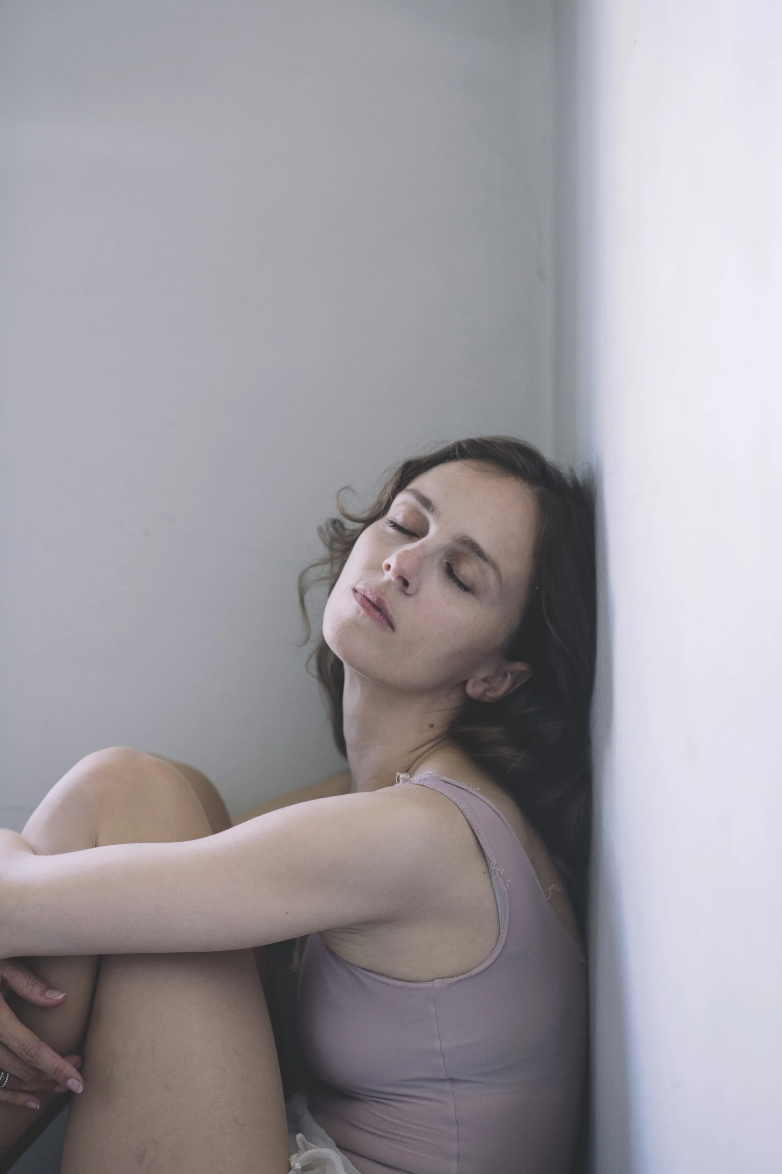Eine junge Frau mit geschlossenen Augen, die im Sitzen an einer weißen Wand lehnt.