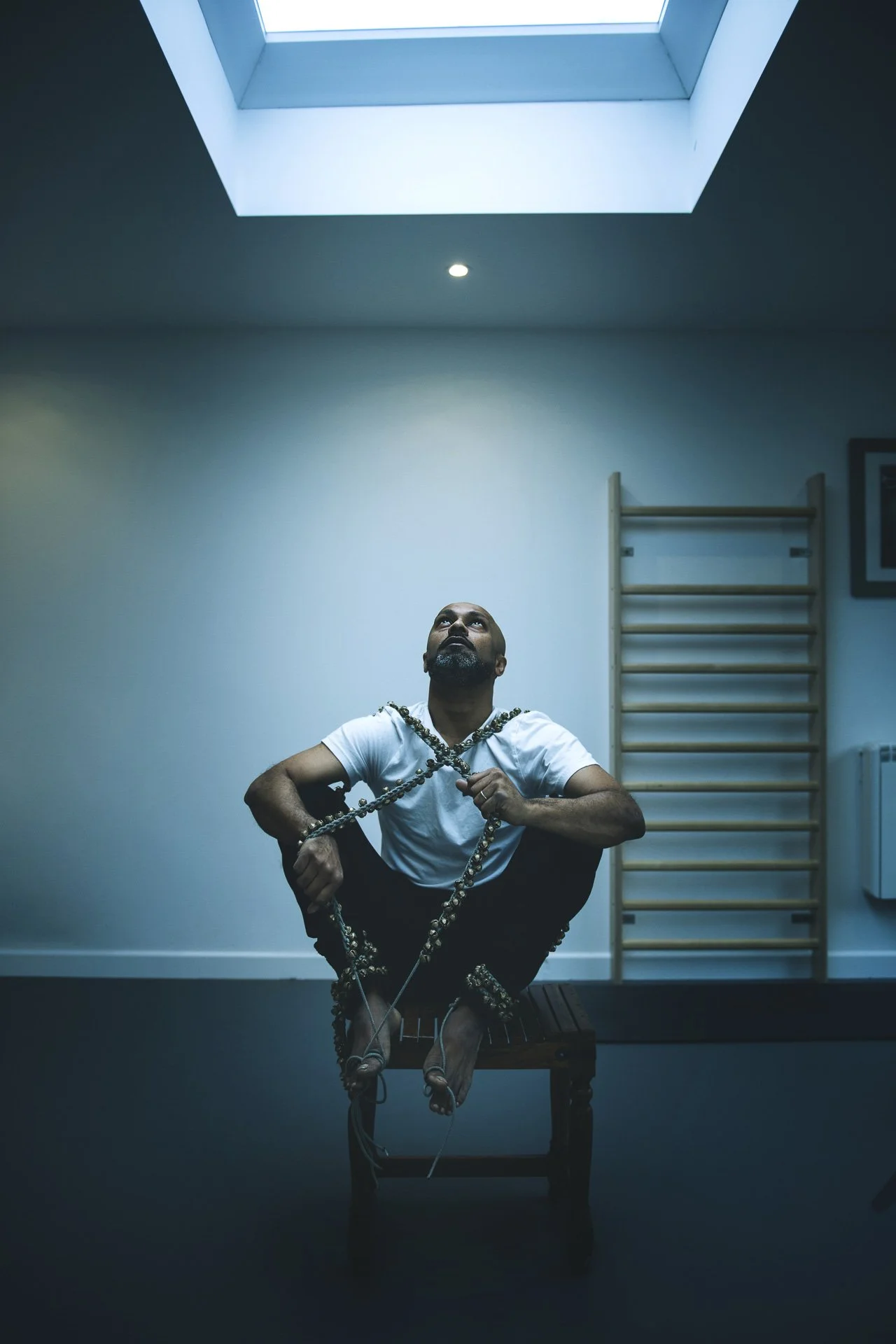 Ein Mann sitzt auf einem Stuhl in einem Raum mit einem Dachfenster. Er hält eine Kette um den Hals, schaut nach oben und hat eine nachdenkliche Haltung.