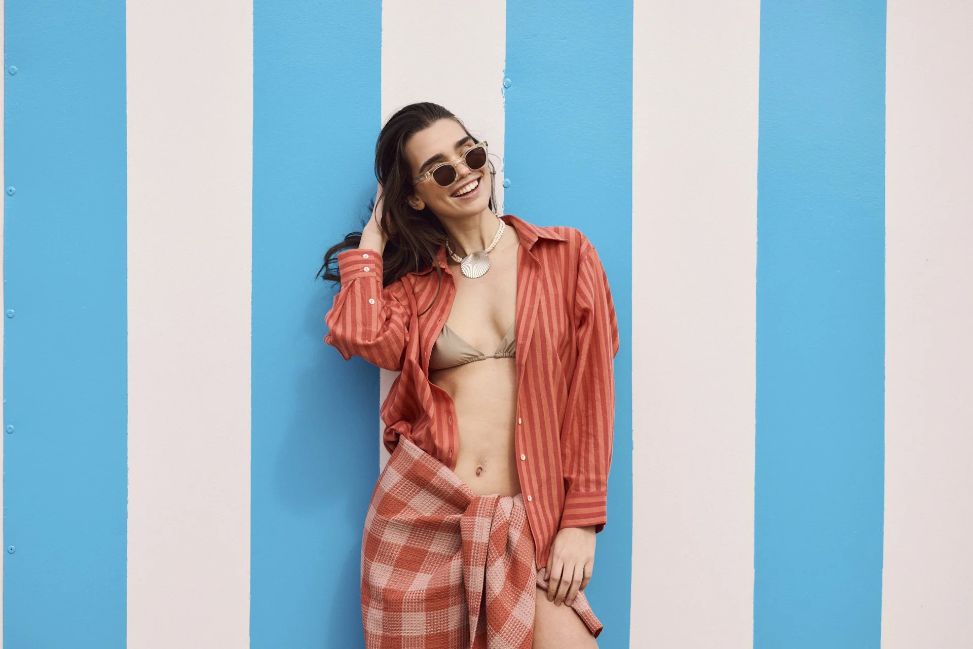 Lächelnde Frau mit Sonnenbrille, rotem Hemd und Bikini-Top steht vor einer blauen und weißen gestreiften Wand.