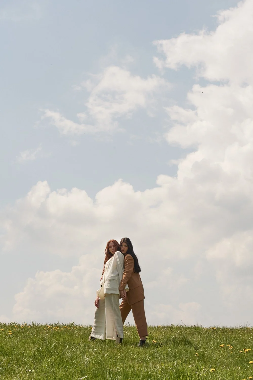 Zwei Frauen stehen auf einer grünen Wiese mit gelben Blumen, im Hintergrund ein Himmel mit Wolken.