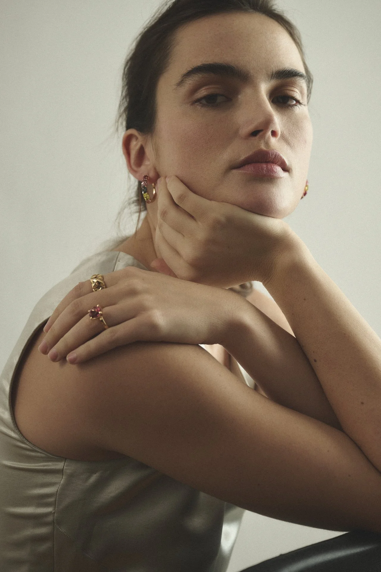 Nahaufnahme eines jungen Mädchens mit dunklen Haaren, das ihre Hand auf ihrem Kinn ruht und Schmuck wie Ringe und Ohrringe trägt, vor einem neutralen Hintergrund.