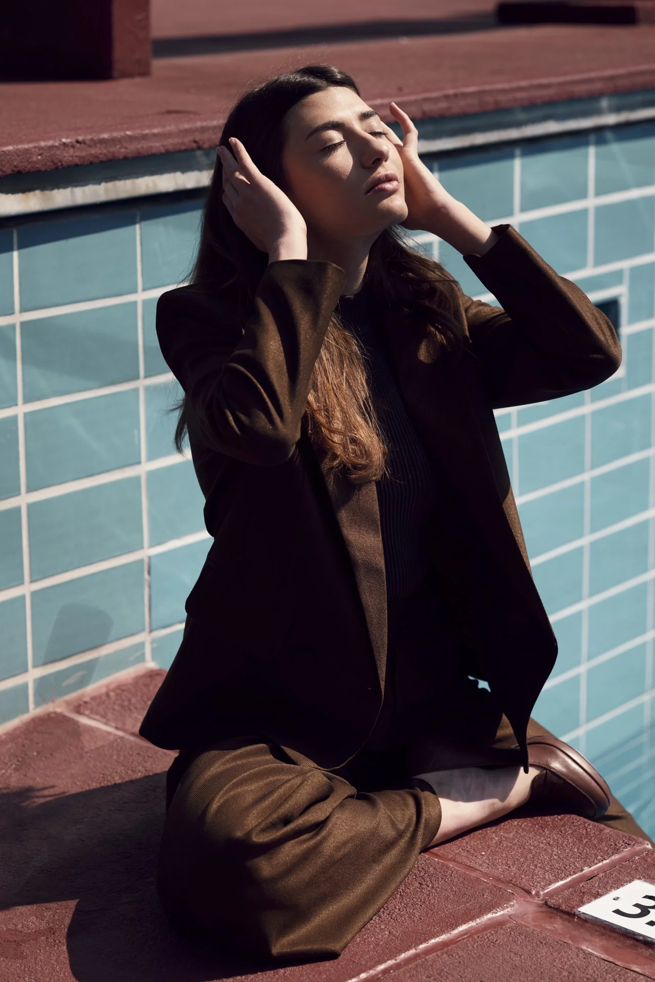 Eine Frau sitzt am Rand eines Schwimmbeckens, hält die Hände an die Seiten ihres Kopfes, die Augen sind geschlossen, und sie wirkt entspannt.