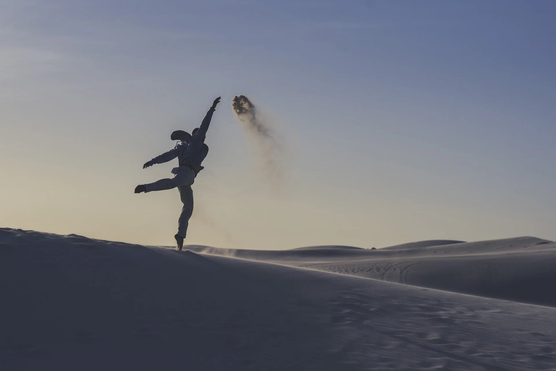 Ein Mensch in Schneeschuhen bei Sonnenuntergang, der einen Handstand auf dem Schnee macht, mit aufsteigendem Staub und dunklem Himmel im Hintergrund.
