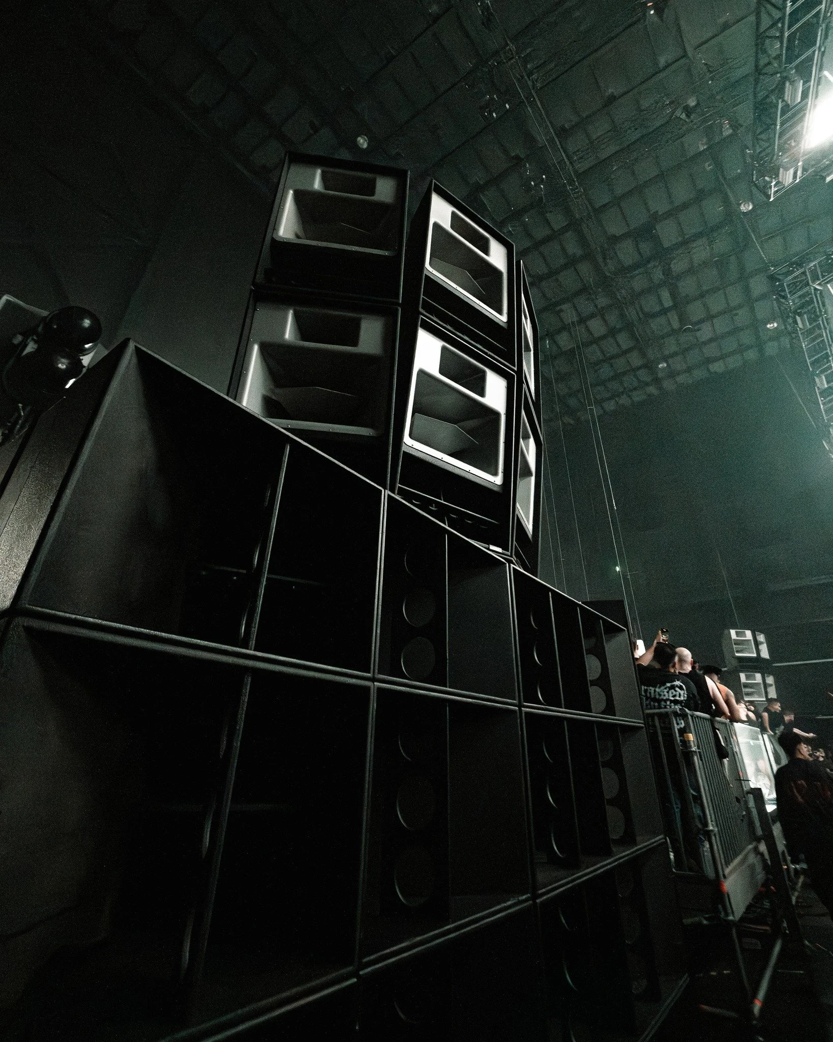 A large stack of black and gray loudspeakers at a concert or event venue with some people in the background.