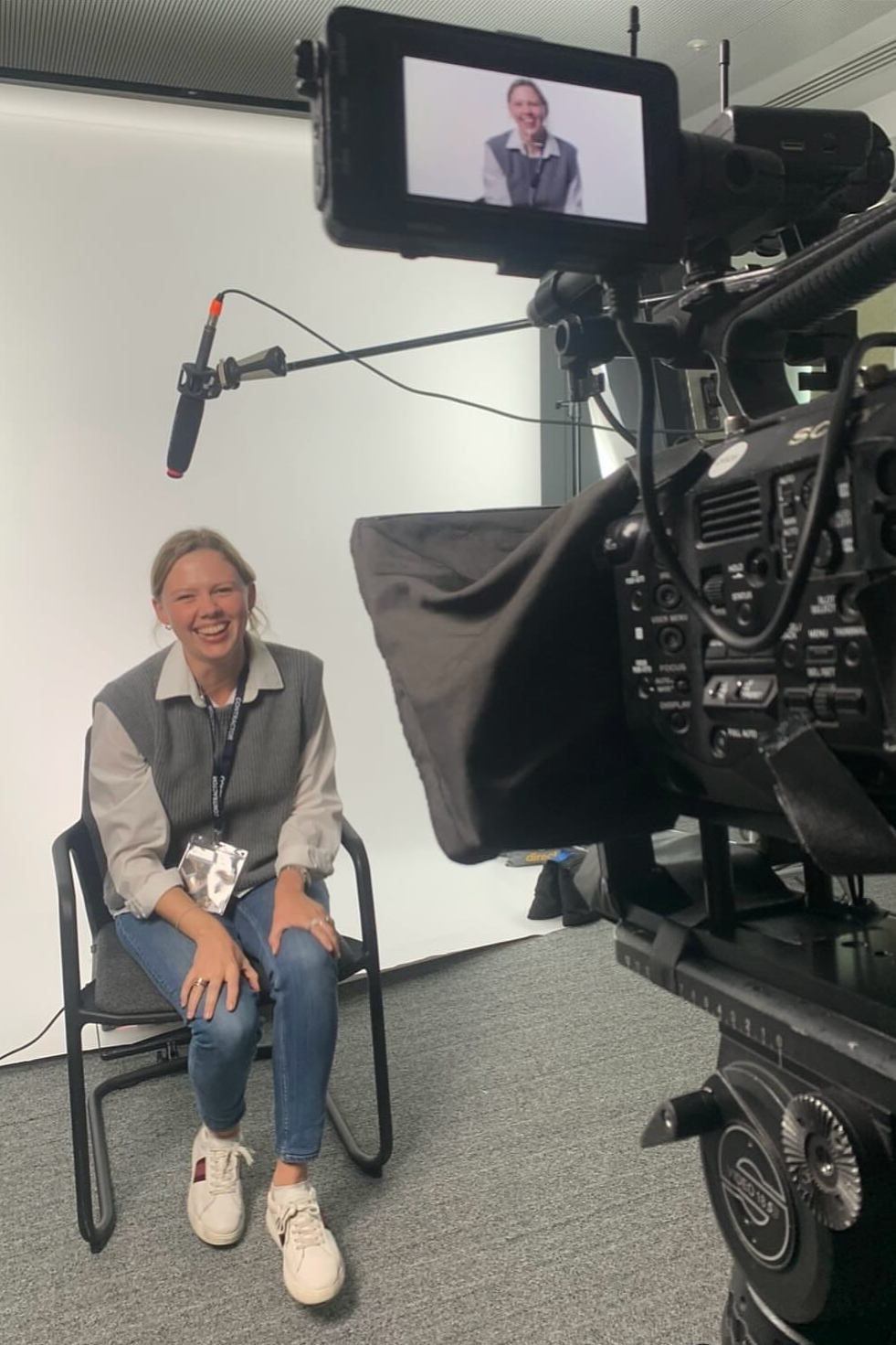 Woman sitting on a chair, laughing during an interview or recording session, with a camera recording her against a plain white background.