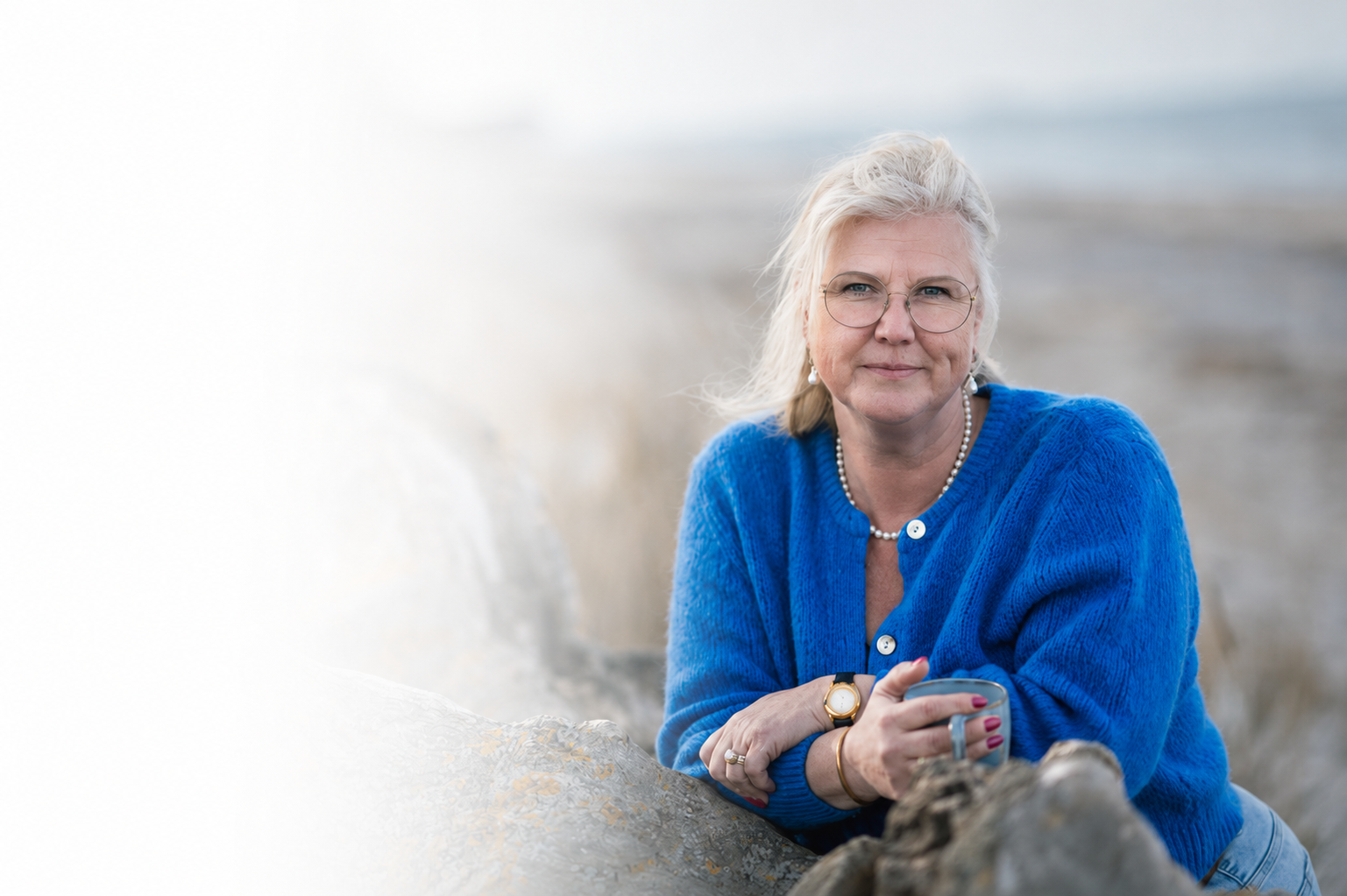 En ældre kvinde med gråt hår, iført en blå sweater, holder en kop ved stranden.