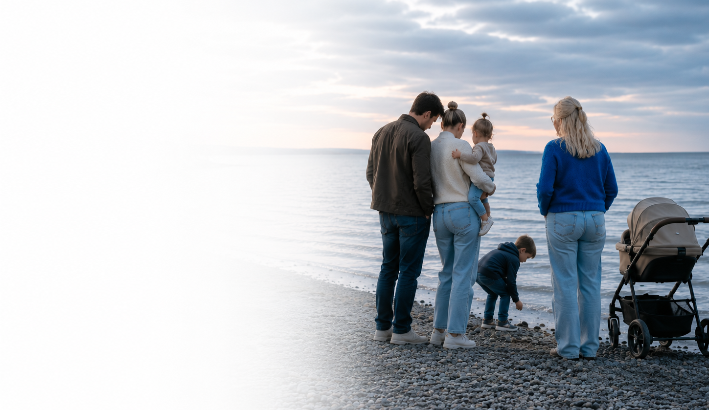 En familie står ved stranden og kigger på vandet, med et barnevogn til højre.