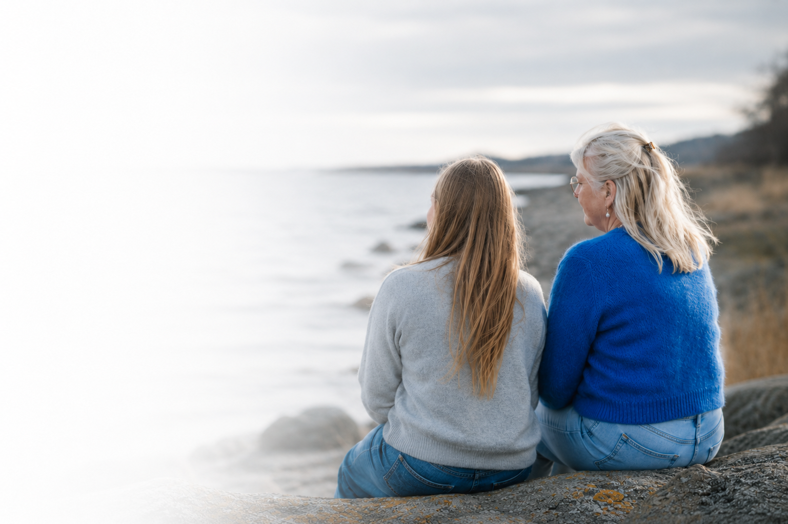 To kvinder sidder på en klippe ved en kyst, kigger ud over havet, bærer afslappede tøj i blå og grå nuancer.