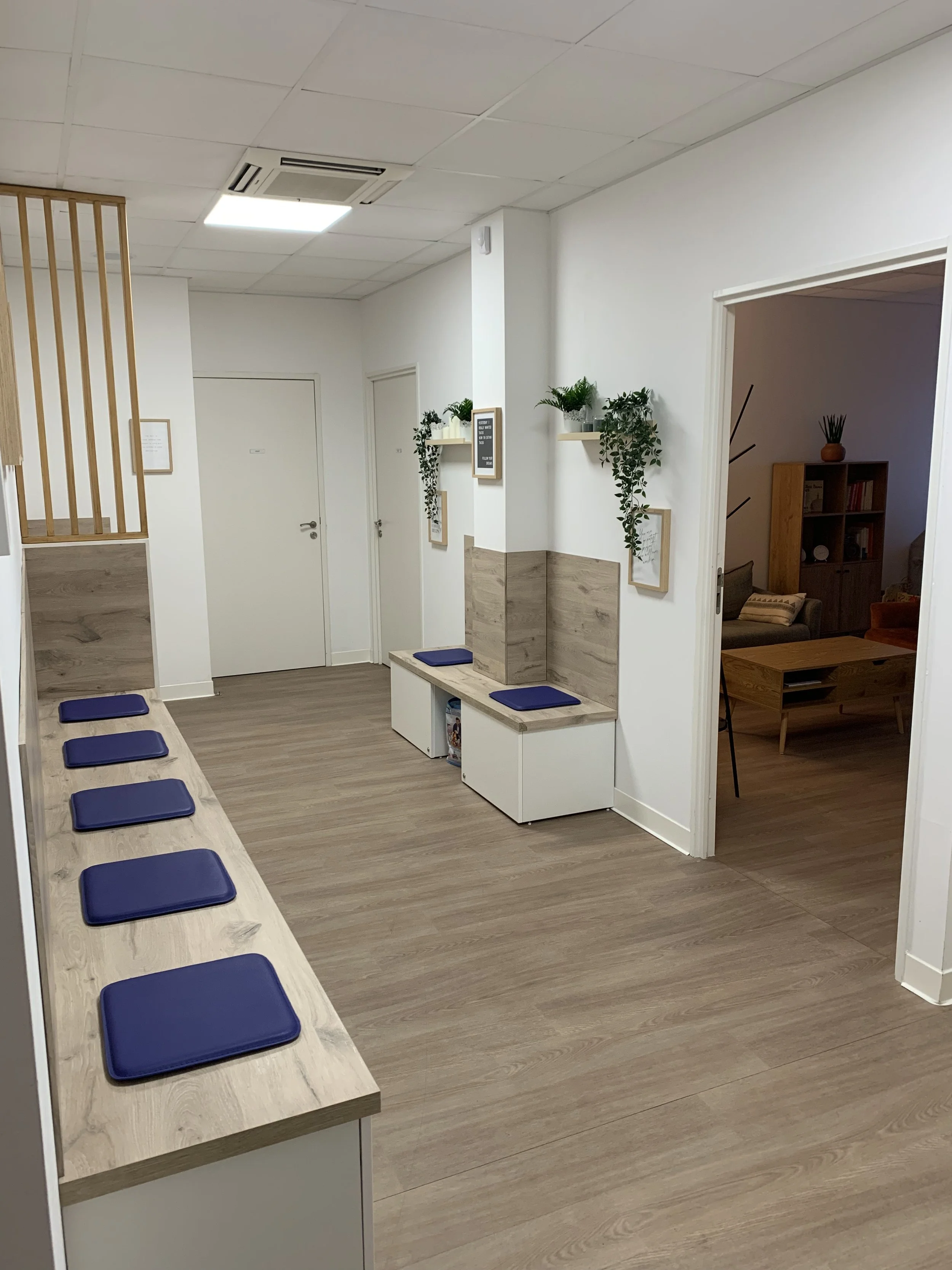 Salle d'attente ou espace commun avec un banc long en bois avec des coussins bleus, murs blancs décorés de plantes en pot et cadres, porte d'entrée en métal blanc, et pièce attenante visible avec mobilier en bois et décoration végétale.