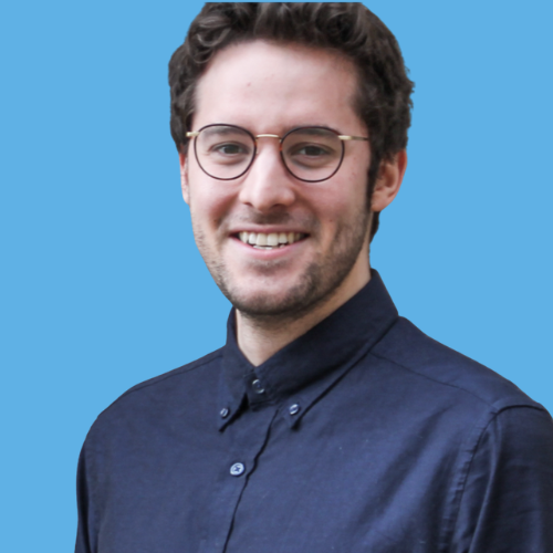 Photo of Micah Kaats, with curly dark hair and glasses, smiling, wearing a dark blue button-up shirt, against a light blue background.