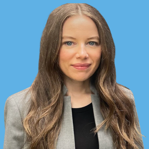 Photo of Dr. Cherise Regier, with long wavy brown hair, blue eyes, wearing a gray blazer over a black top, standing against a solid blue background.