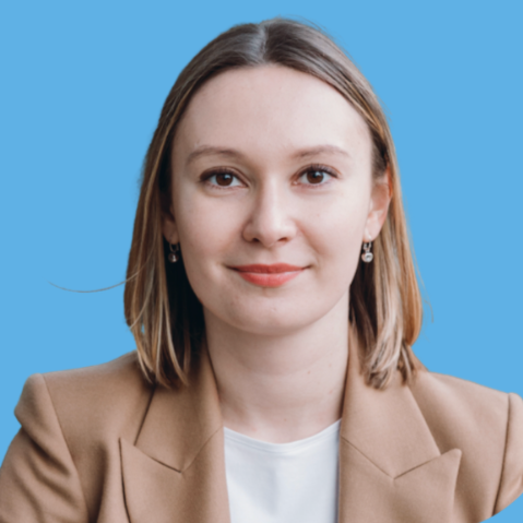 Photo of Dr. Alexandra Kirienko, with straight light brown hair, wearing a tan blazer and white shirt, smiling at the camera, against a blue background.