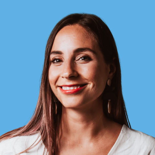 Photo of Janeane Tolomeo, close-up portrait of a woman with long brown hair wearing a white top and earrings, smiling against a solid blue background.