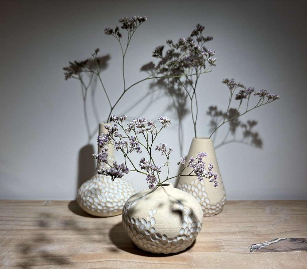Trois vases en céramique blanche avec des motifs sculptés, contenant des branches de fleurs violettes, sur une surface en bois.