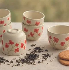 Tasses et bols en céramique blancs avec des motifs de fleurs de cerisier, quelques feuilles de thé et biscuits sur une table.