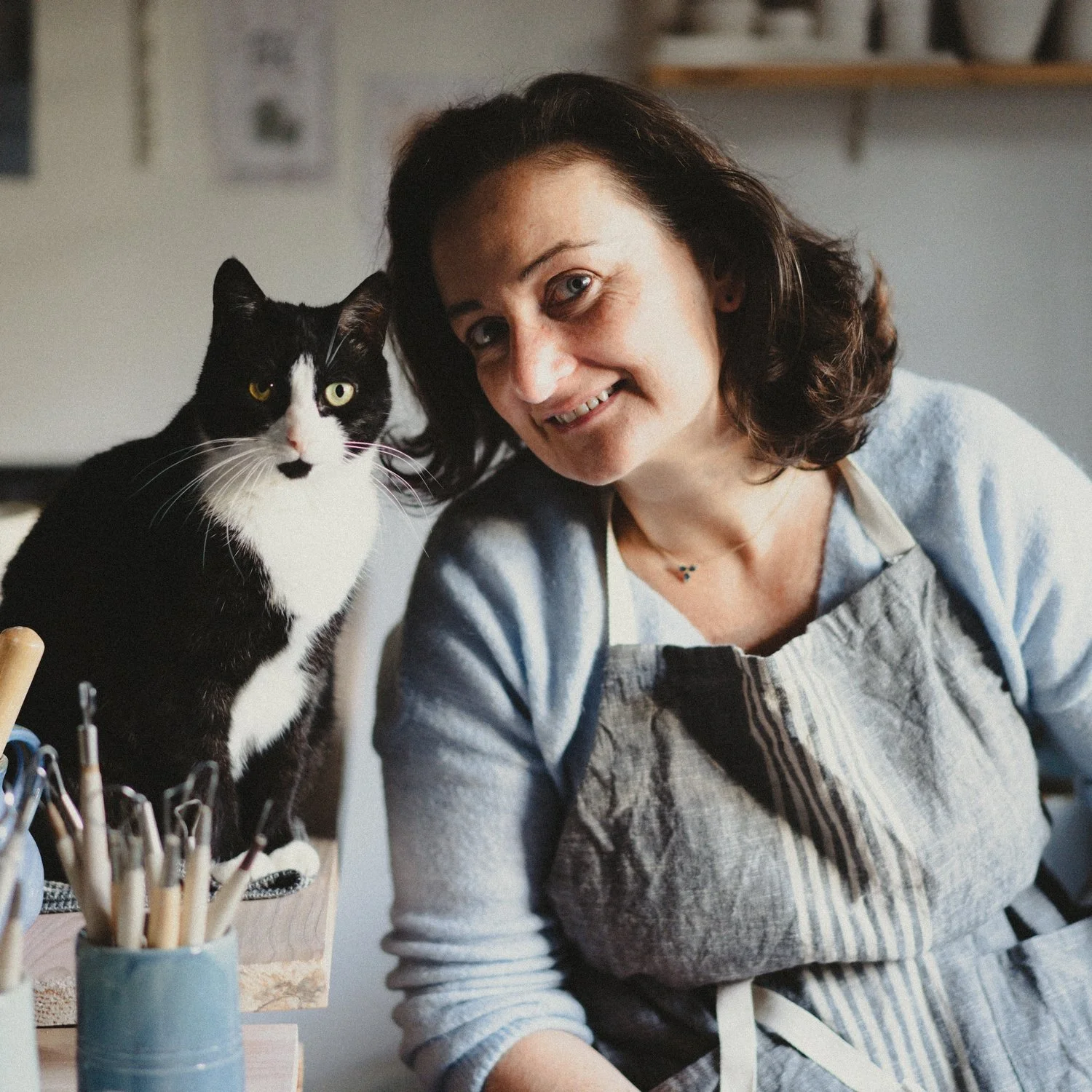 Artisan potière souriante en tablier, assise dans son atelier avec un chat noir et blanc à ses côtés.