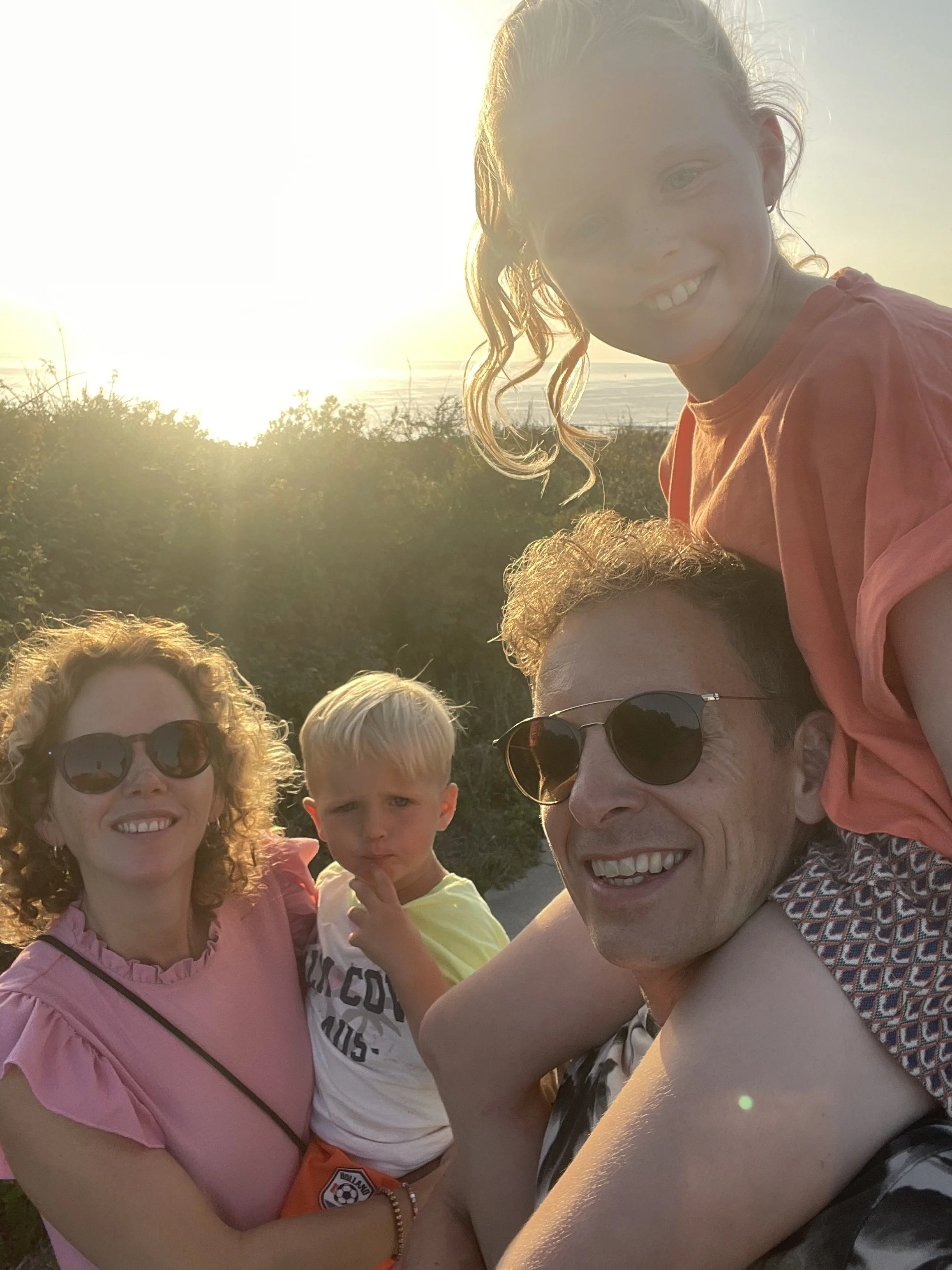Groep mensen, inclusief kinderen, maakt een selfie tijdens zonsondergang op het strand.