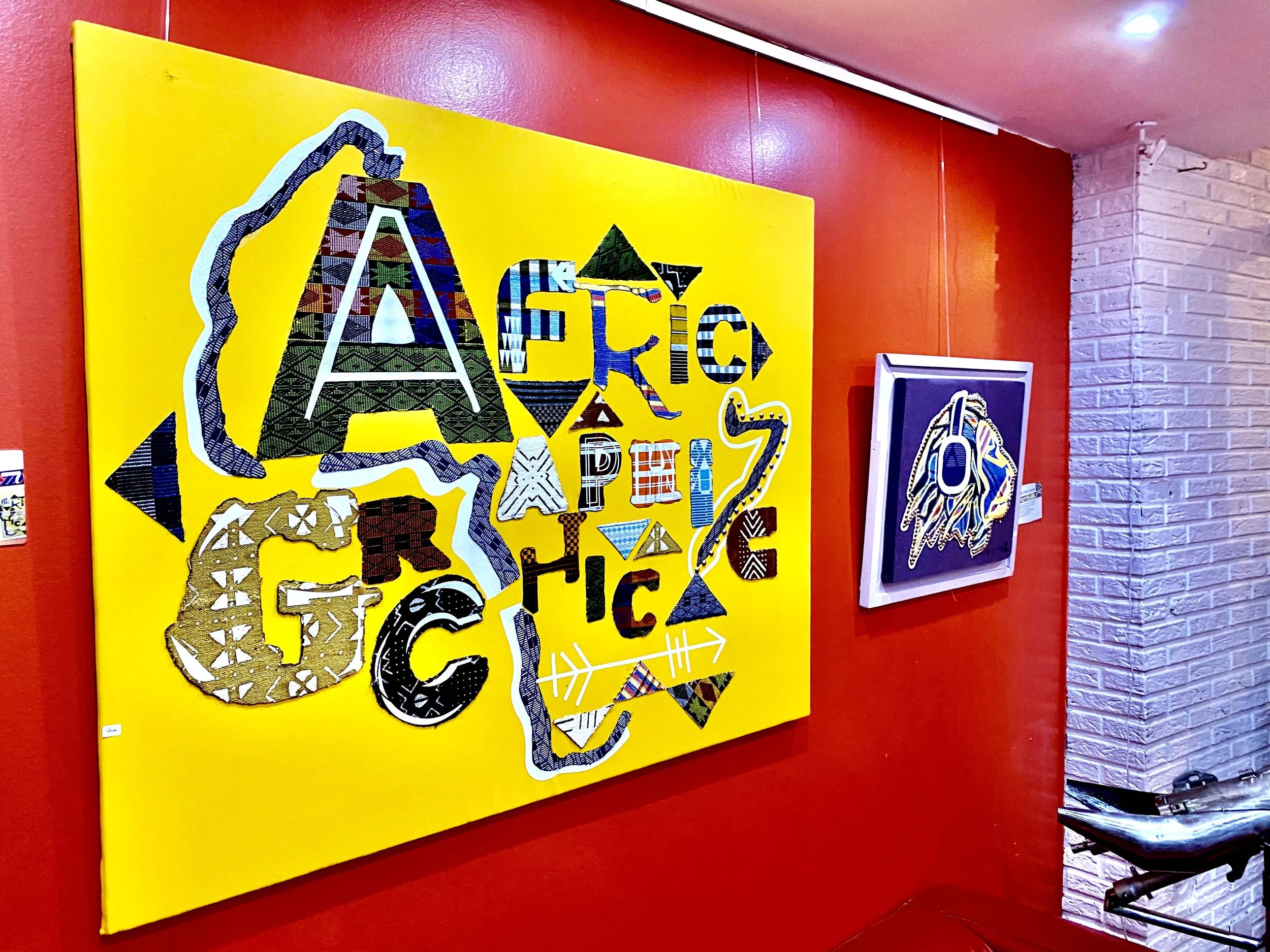 Exposition d'œuvres d'art abstraites avec lettres et motifs colorés sur un fond jaune et encadrée en blanc, dans une galerie avec murs rouge et blanc
