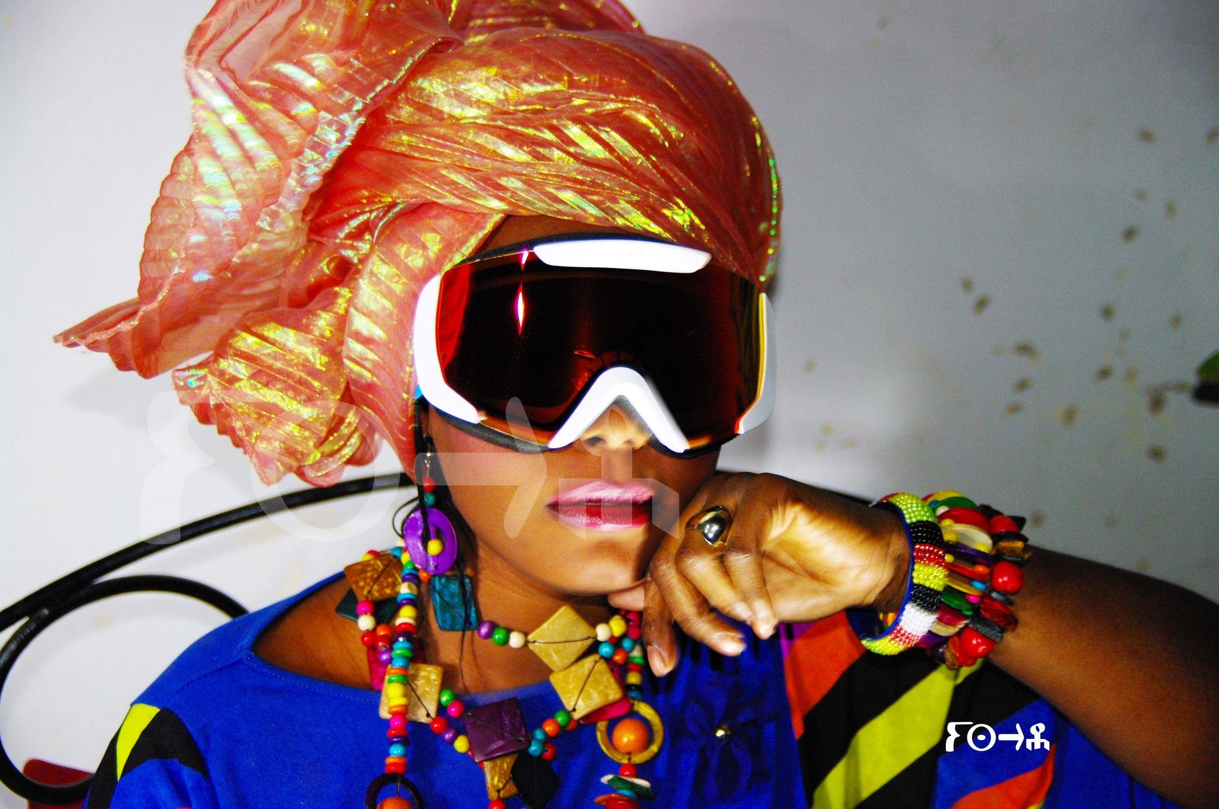 Une femme portant un turban coloré et des lunettes de ski noires réfléchissantes. Elle est décorée avec plusieurs bijoux colorés et porte une tenue vibrante, avec un fond simple et peu détaillé.Afa Faty Art Numérique - Matrice digitale Full HD