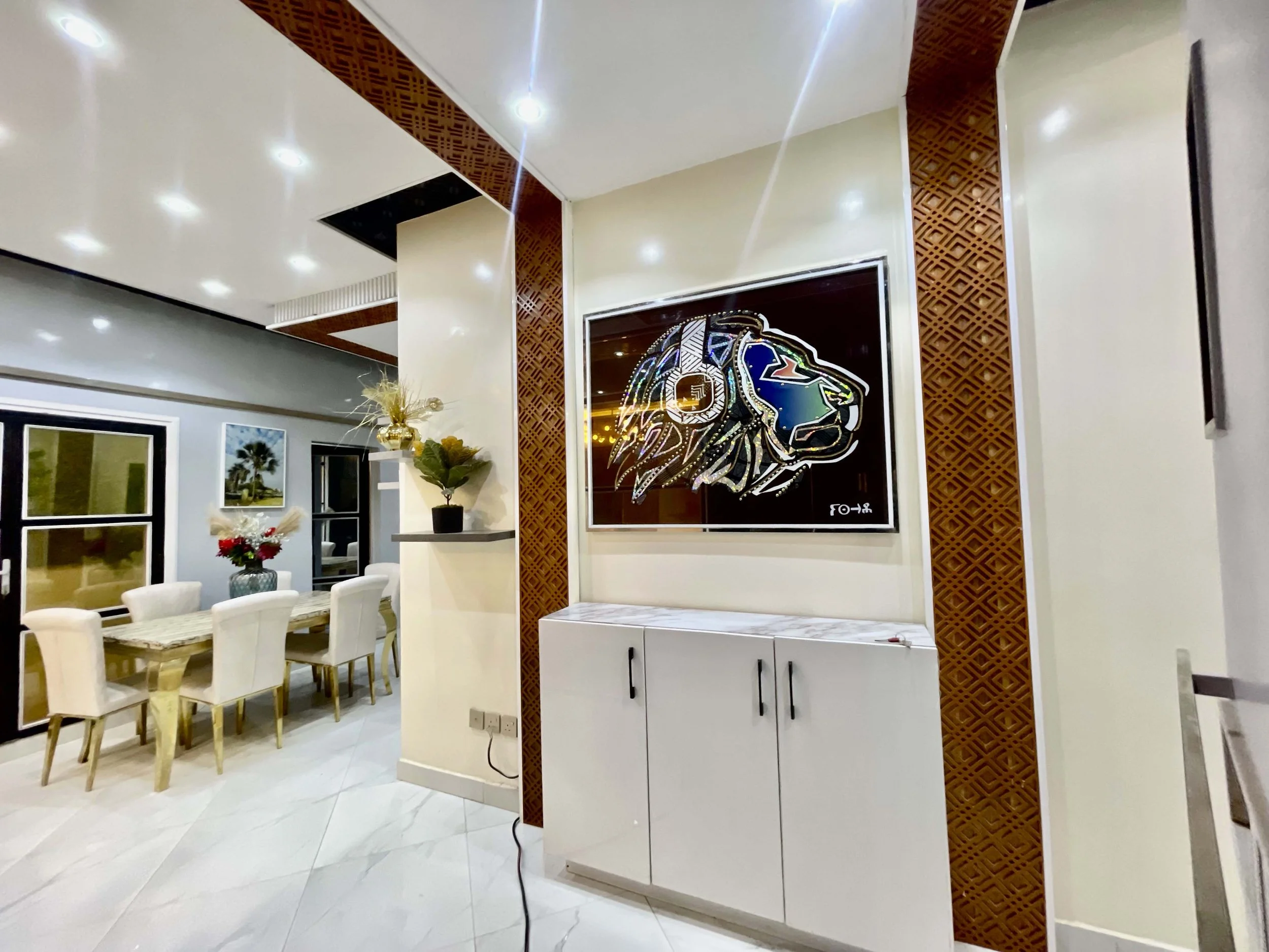 Intérieur moderne avec un mur décoratif, une image d'un lion stylisé en métal, une armoire blanche, une table de salle à manger avec des chaises et quelques plantes décoratives.