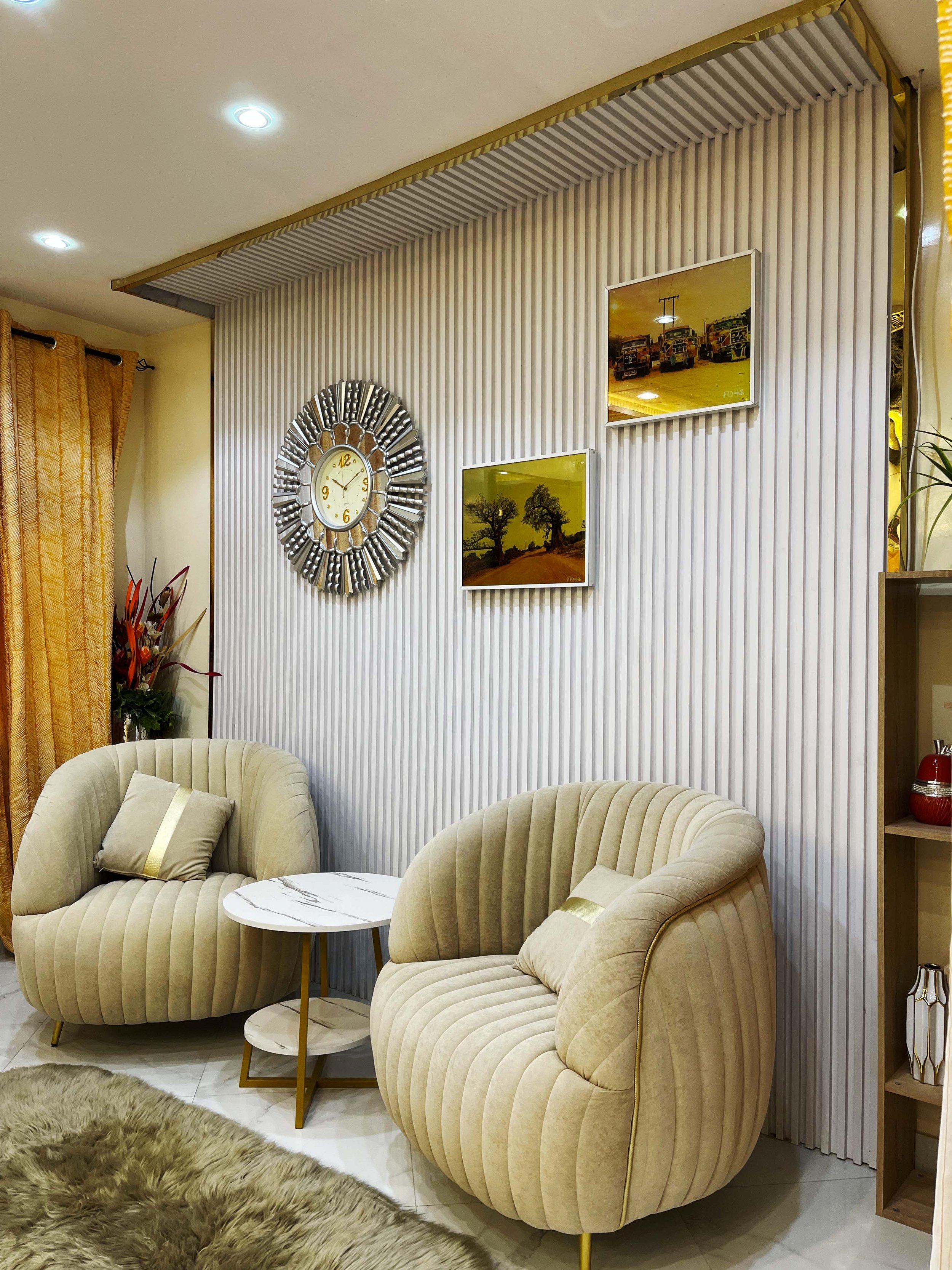 Salon avec deux fauteuils en velours beige, tableau, horloge design, rideaux dorés, lampe, tapis en fourrure, étagère avec décoraciones.