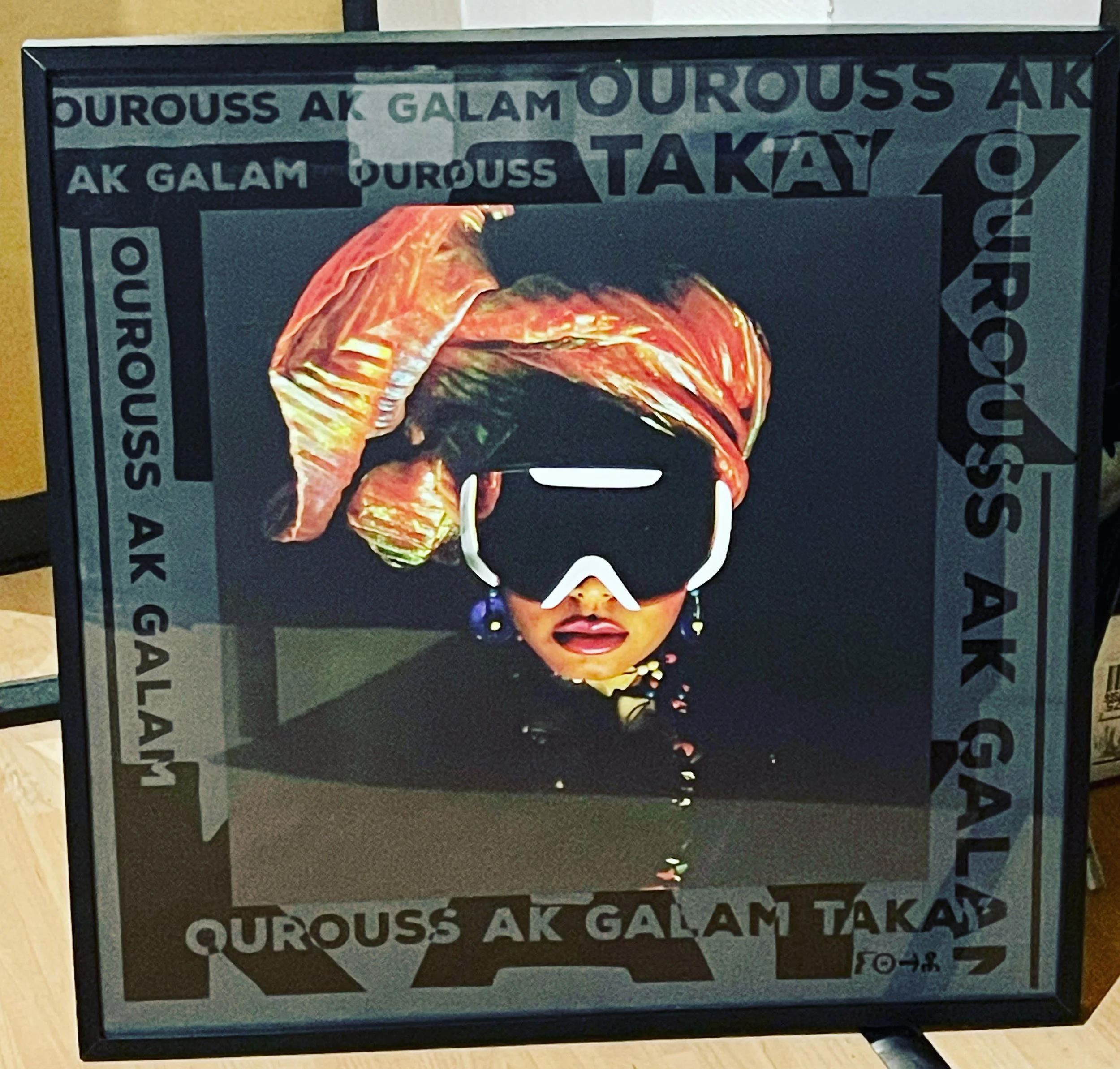 Une personne portant un turban coloré, des lunettes de plongée, des boucles d'oreilles, un collier, maquillée avec du rouge à lèvres foncé, regarde vers l'avant avec un fond sombre. Texte en marges: 'Galam Qurous Ak Takay'. Afa Faty art for artefacts