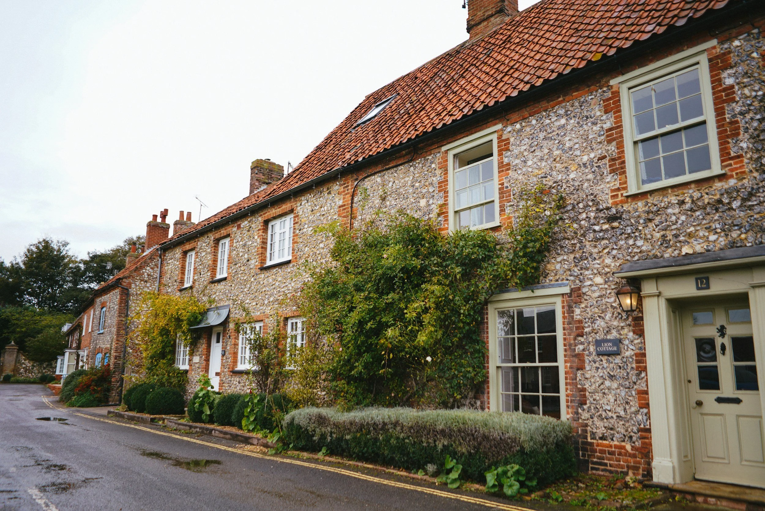 A row of brick and flint townhouses with red tiled roofs, white framed windows, and front gardens with bushes and trees, on a wet street in an urban neighborhood.