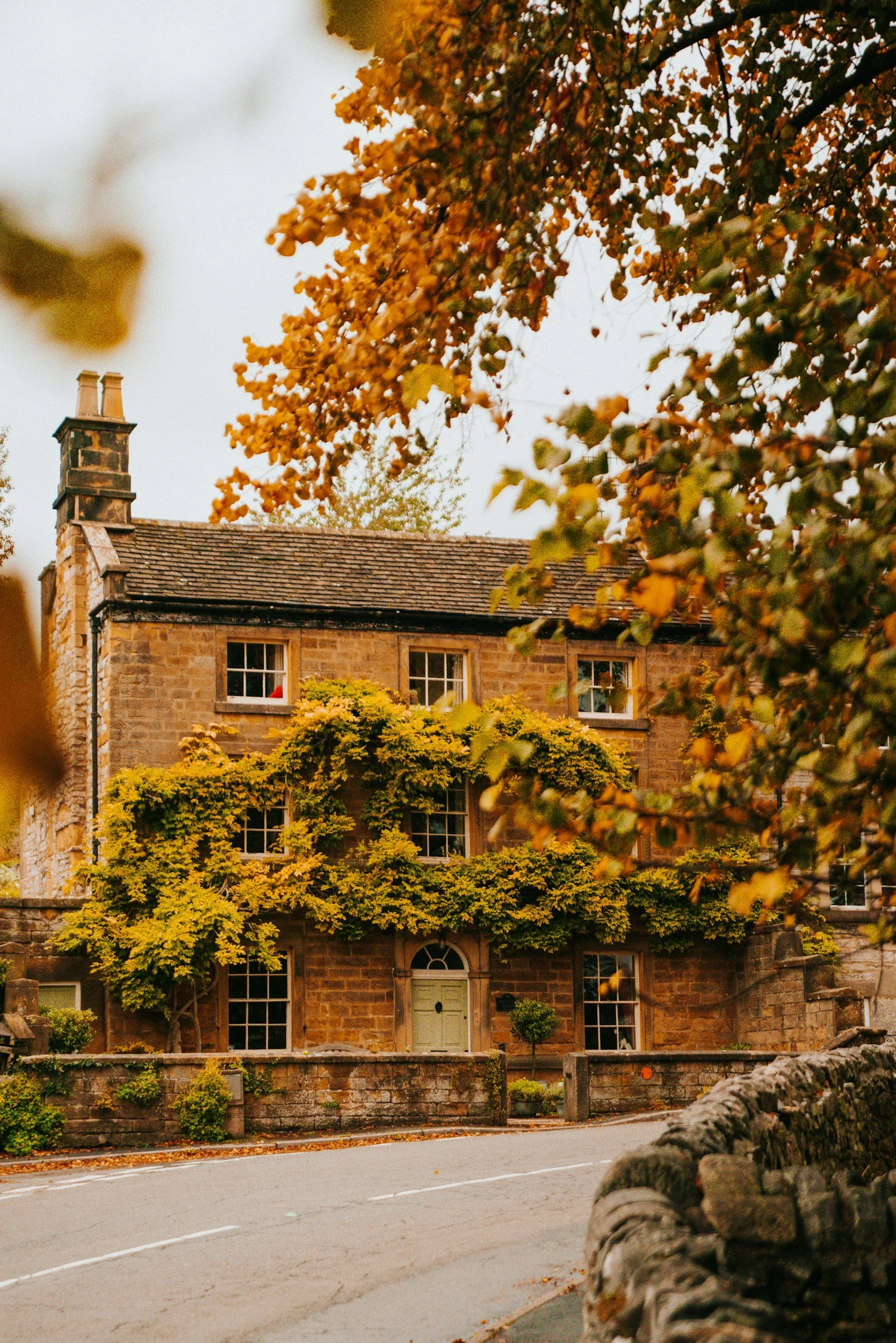 A stone house with green door and multiple windows, partially covered with autumn foliage, located beside a curved paved road.