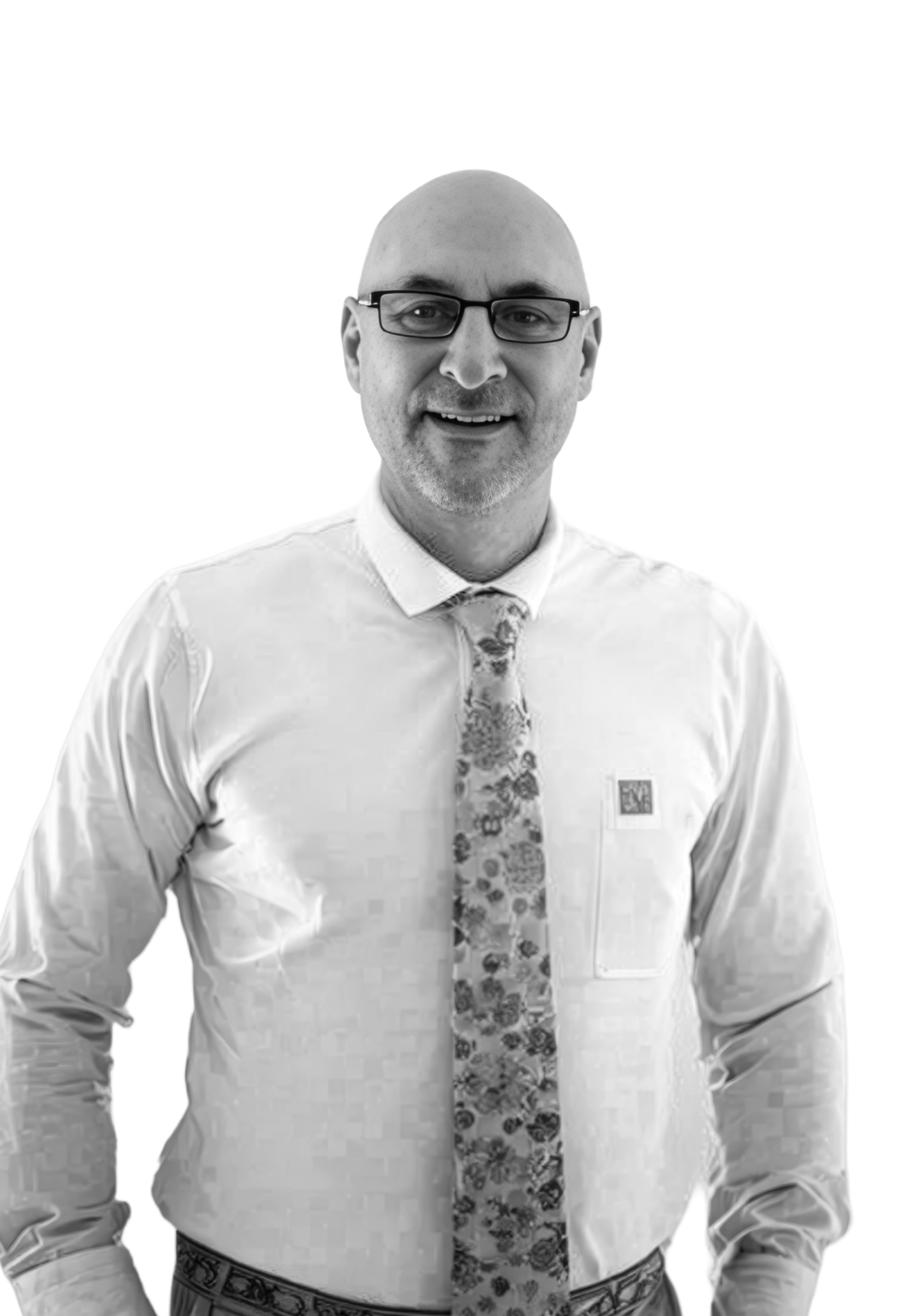 Black-and-white photo of a bald man wearing glasses, a white dress shirt, a patterned tie, and patterned pants, standing against a plain white background.