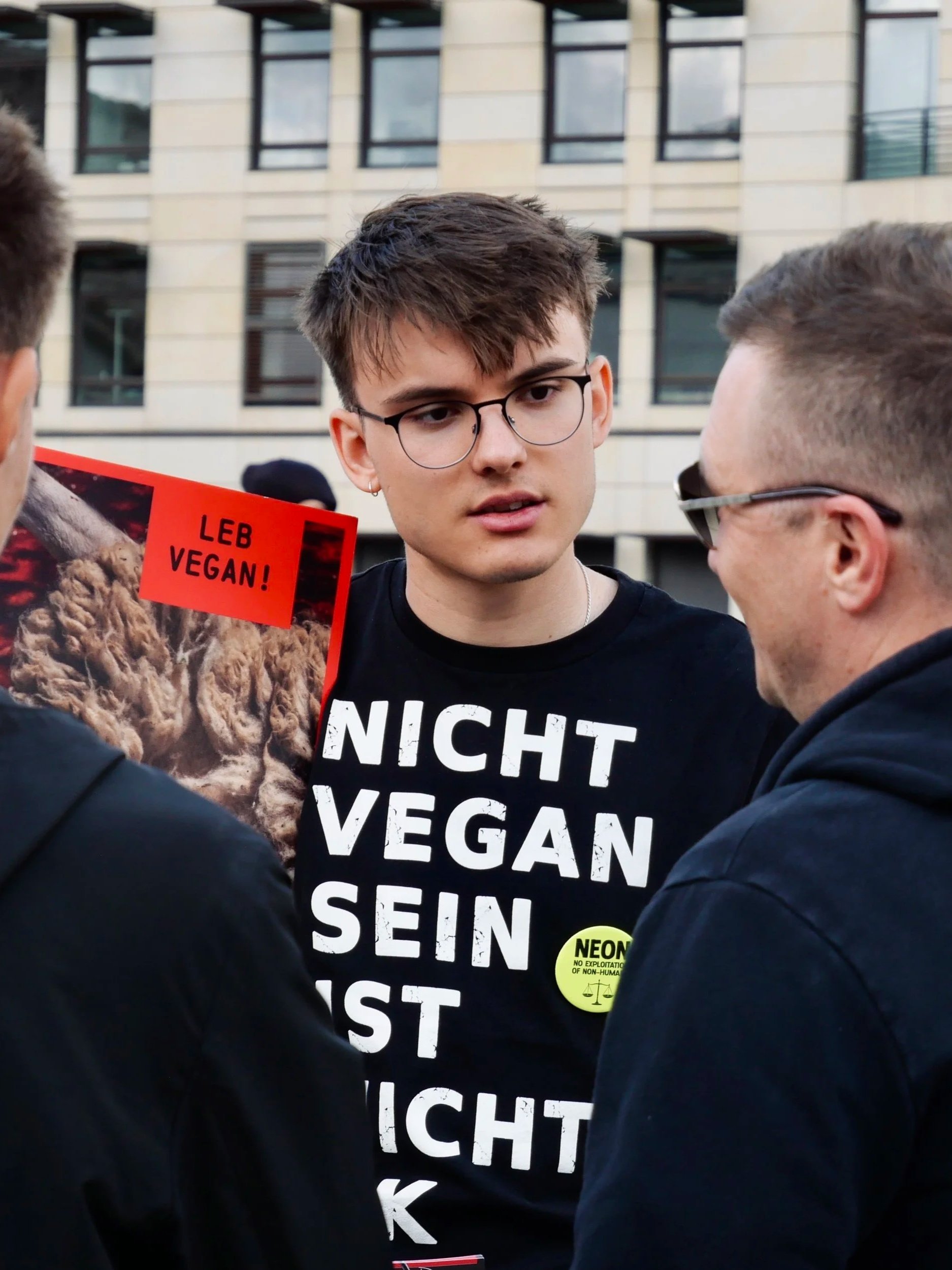 Junge trägt ein schwarzes T-Shirt mit weißen Texten und einem gelben Sticker, spricht mit anderen Personen, im Hintergrund ein modern gebautes Gebäude.