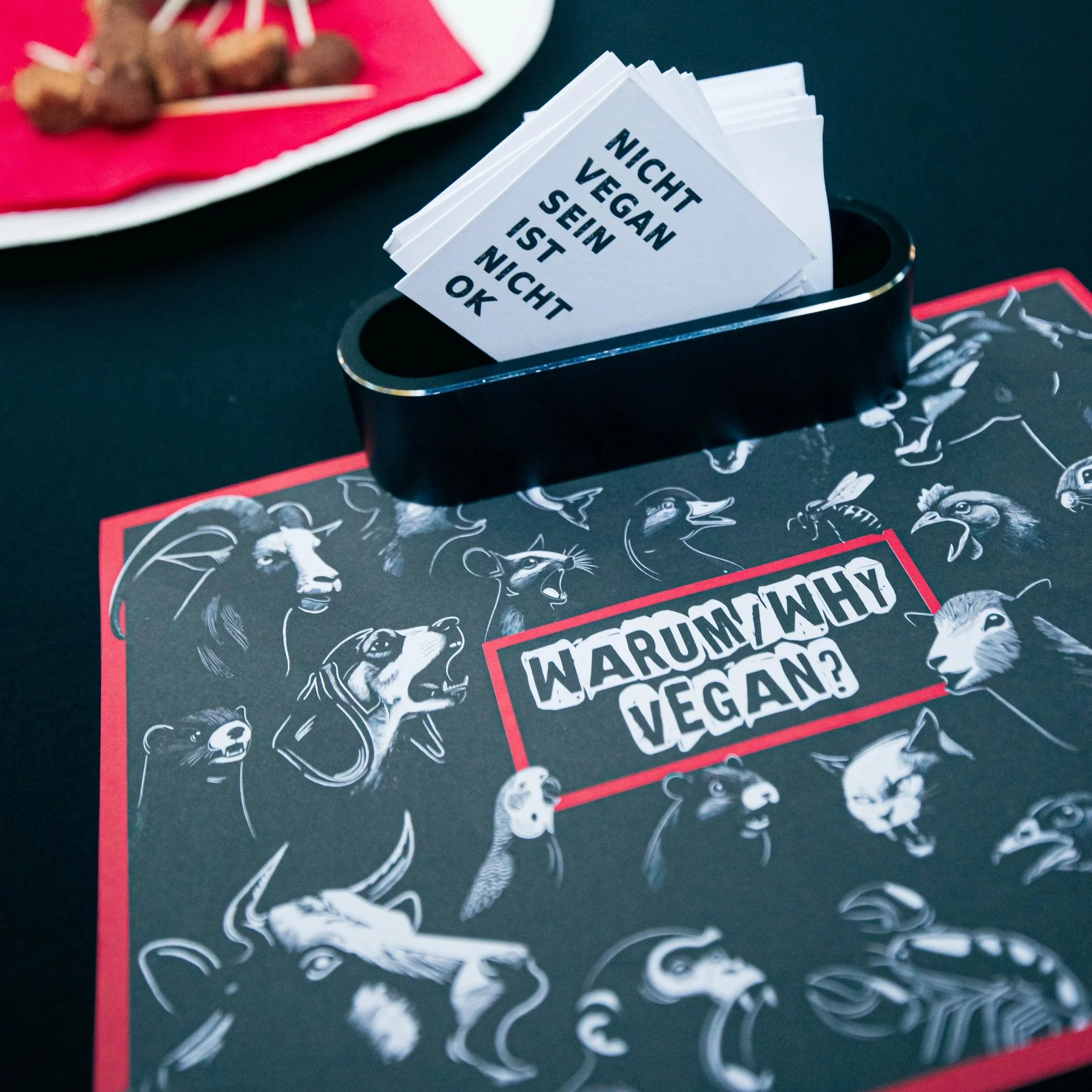 Schwarzes Brett mit weißen Tierzeichnungen und rotem Rahmen, darauf liegt eine Karte mit schwarzer Schrift, die sagt: "Nicht vegane sein, weil es nicht vegan ist, okay"