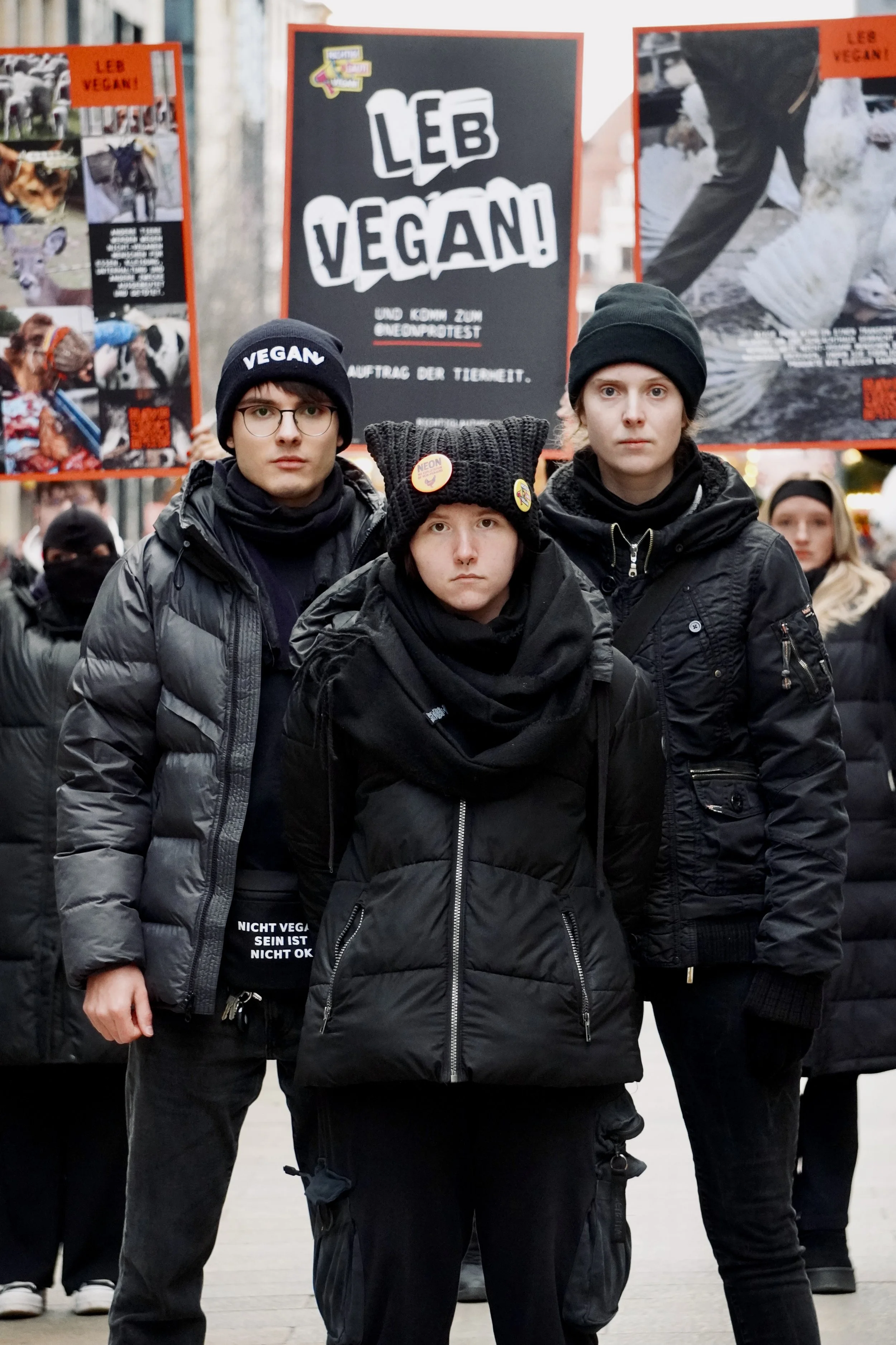 Drei junge Menschen stehen in Schwarz gekleidet in einer Menschenmenge, im Hintergrund ein Plakat mit der Aufschrift 'Leb vegan!'.