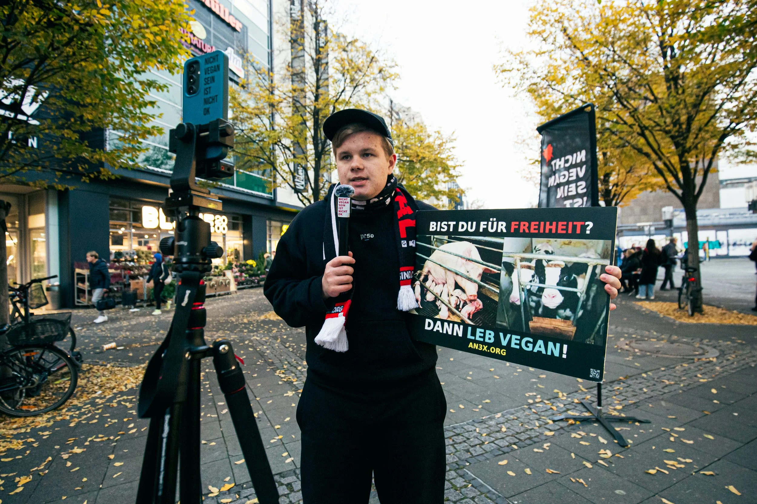 Ein junger Mann hält ein Schild bei einer Demonstration in einer Stadt. Das Schild fordert die Freiheit und zeigt Bilder von Tieren. Im Hintergrund sind Geschäfte und Baumbänke zu sehen, mit herabgefallenen Blättern auf dem Boden.