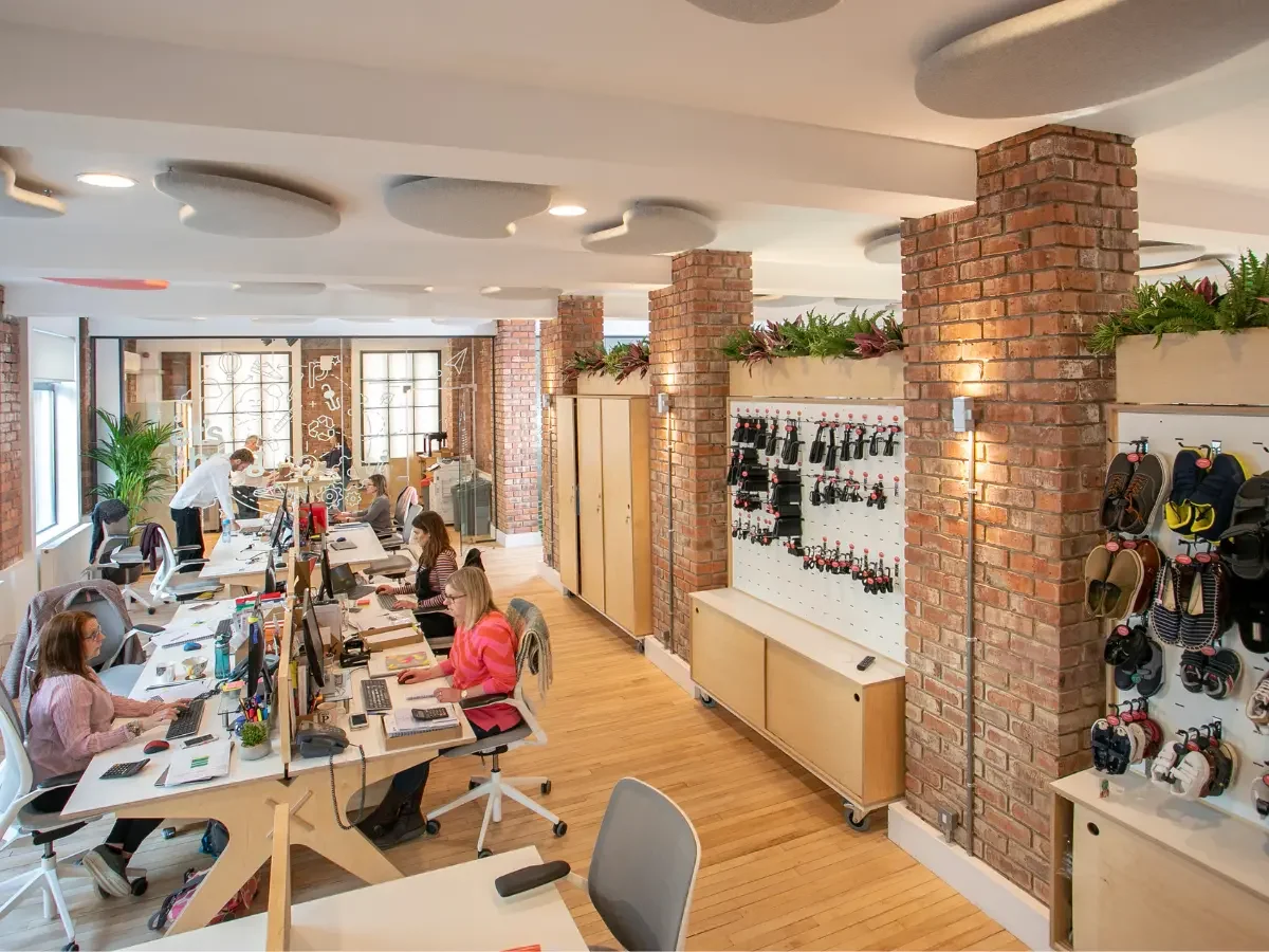 Modern open-concept office space with exposed brick pillars, wooden floors, multiple employees working at desks with computers, and storage areas with tools and accessories on pegboards.