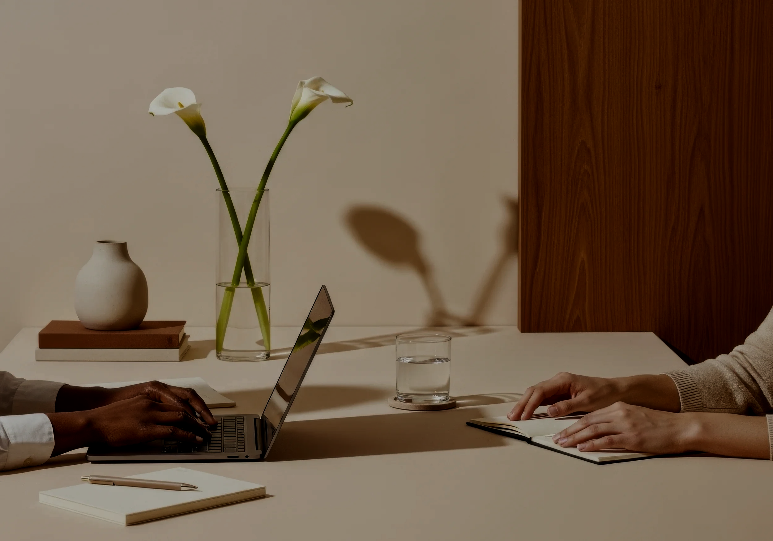 Two people working on laptops at a desk, with a vase of calla lilies and a glass of water in the background.