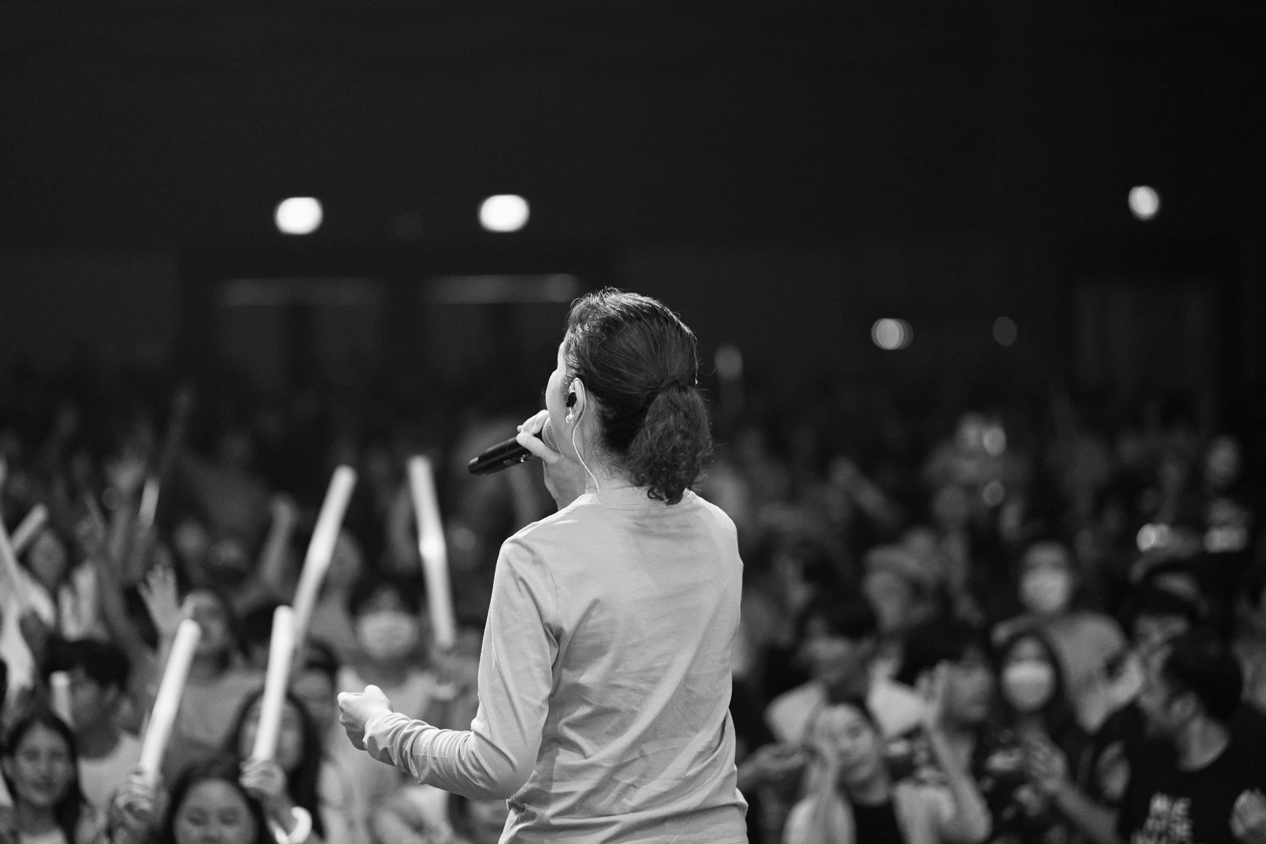 A woman with dark hair tied back, wearing earbuds and a long-sleeve shirt, speaking into a microphone on stage in front of a large seated audience at an indoor event.
