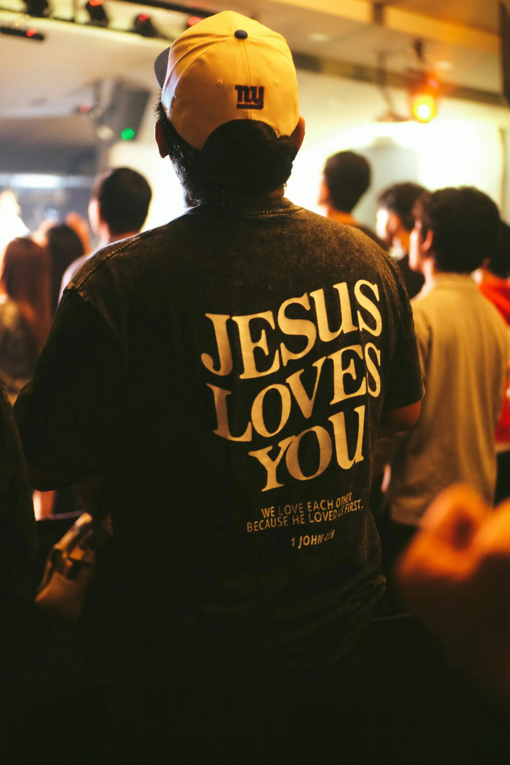 Person wearing a black jacket with 'JESUS LOVES YOU' written on the back, in a crowd at an indoor event, wearing a white cap with an NFL logo.