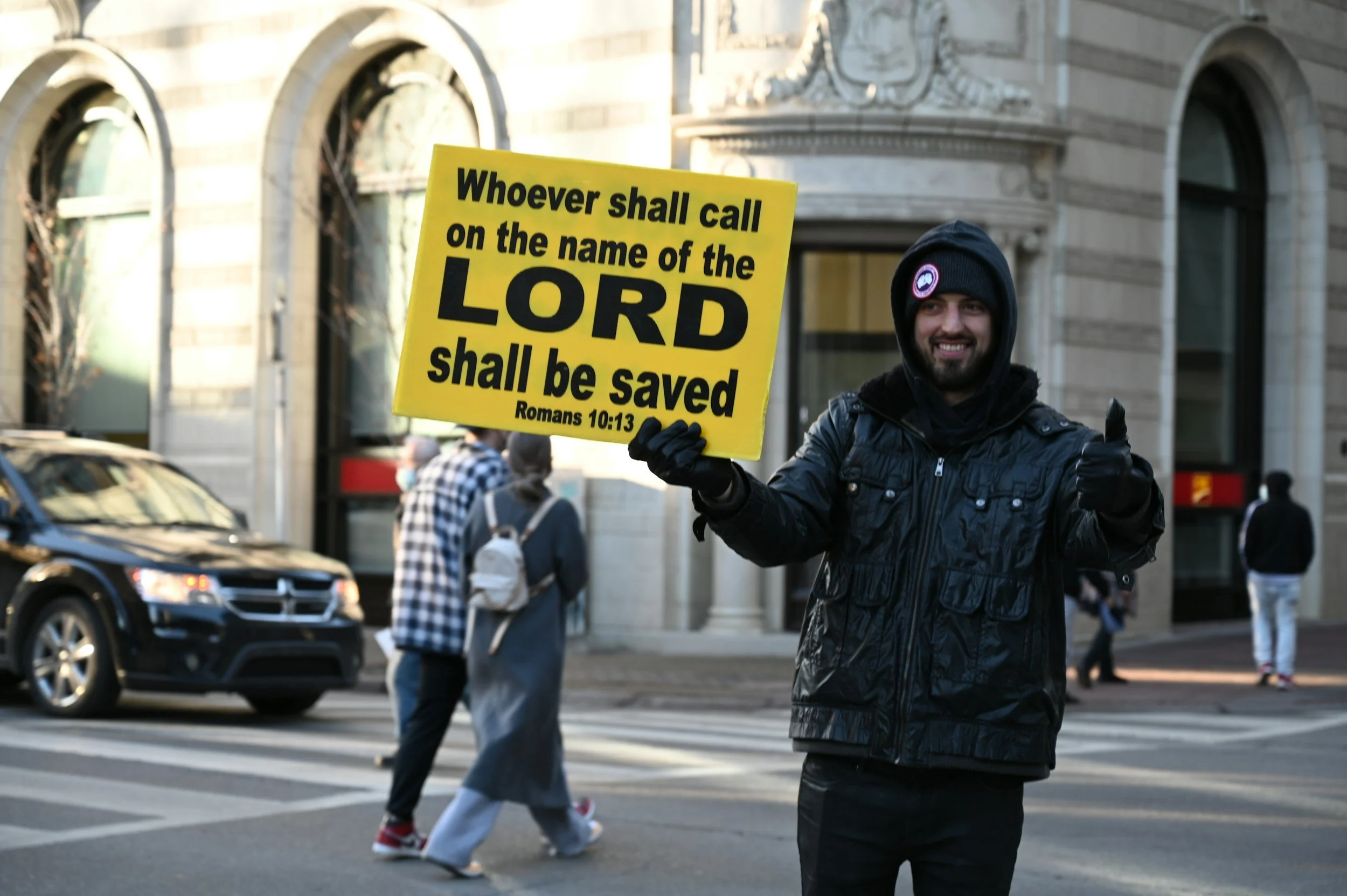 Man wearing black jacket and hoodie holding a yellow protest sign with a Bible quote in an urban street scene.