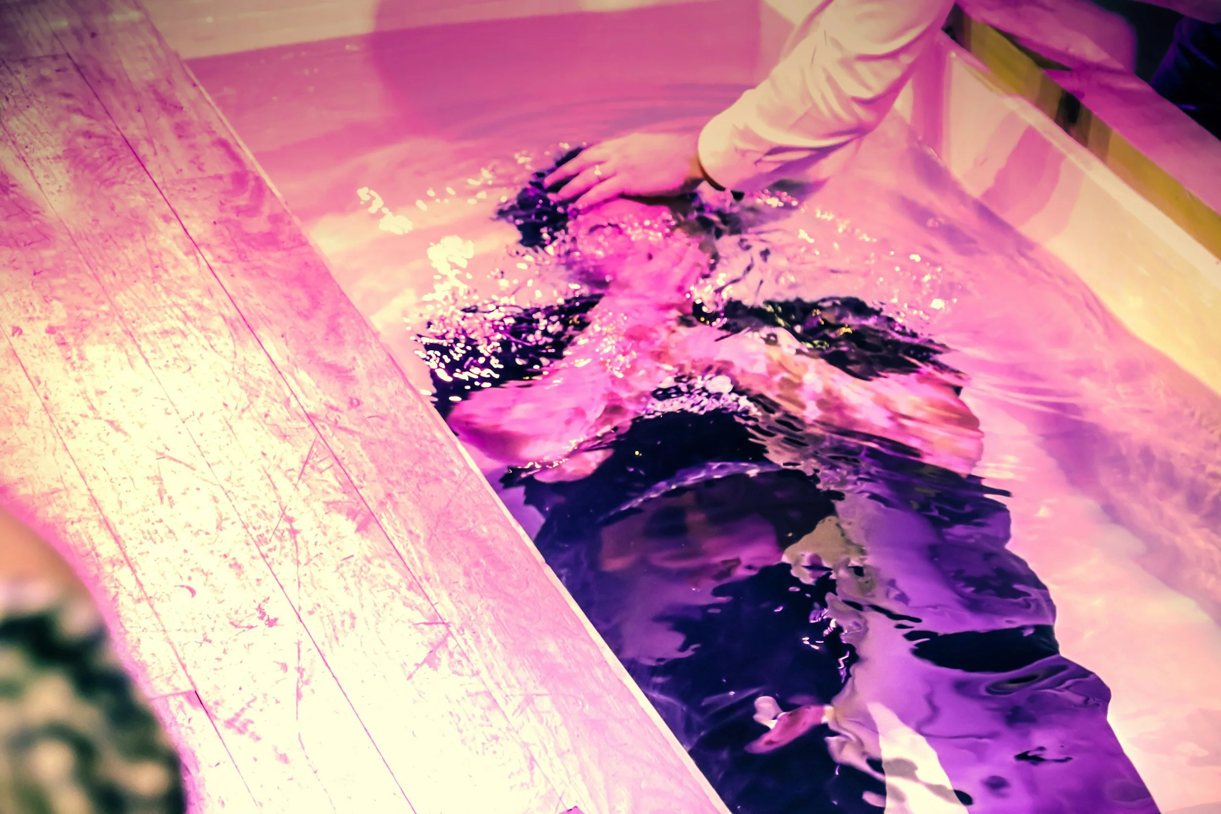 A person in business attire performing a baptism by immersion in a baptismal pool with pink lighting.