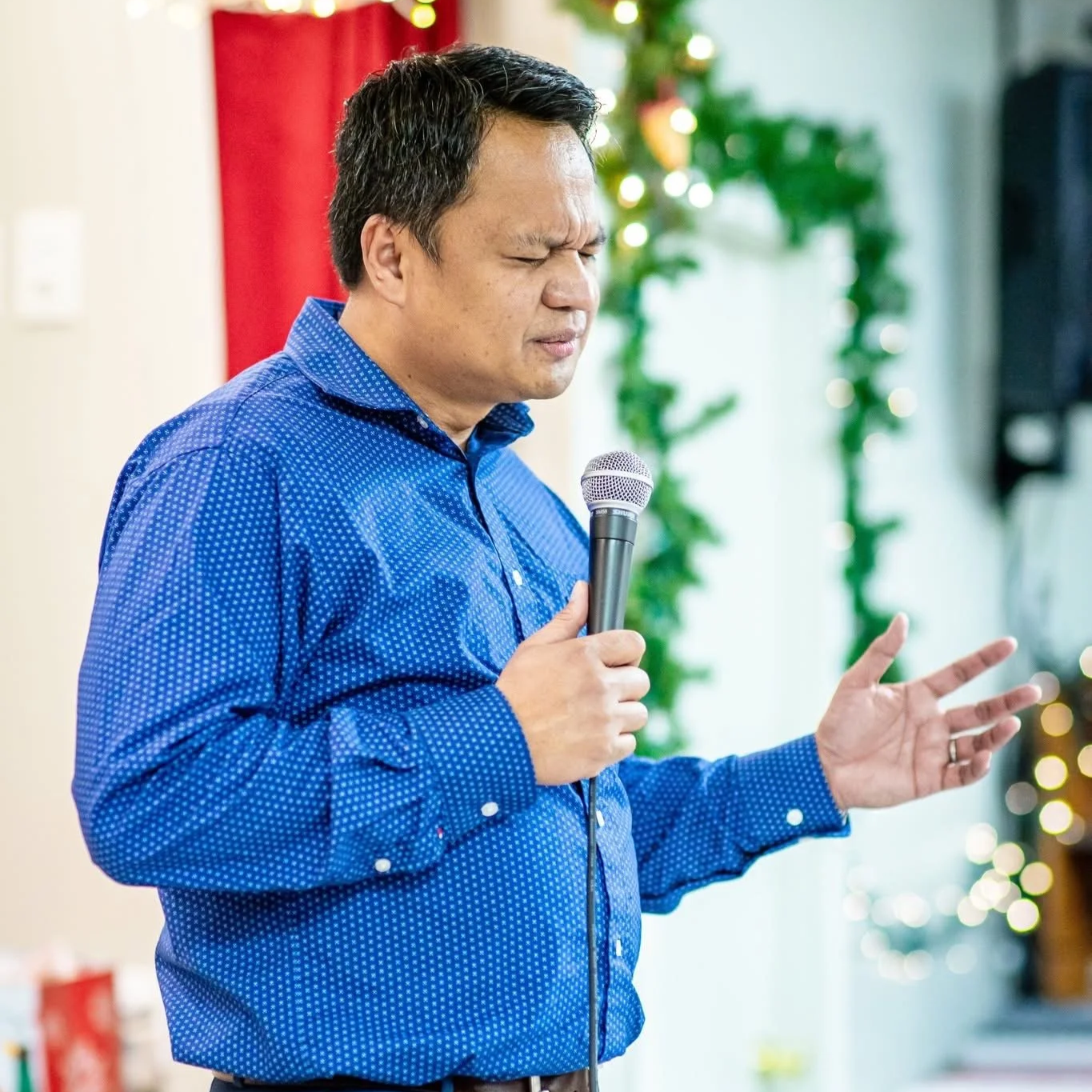 A man in a blue patterned dress shirt holding a microphone with his eyes closed and one hand raised, standing indoors decorated for Christmas.