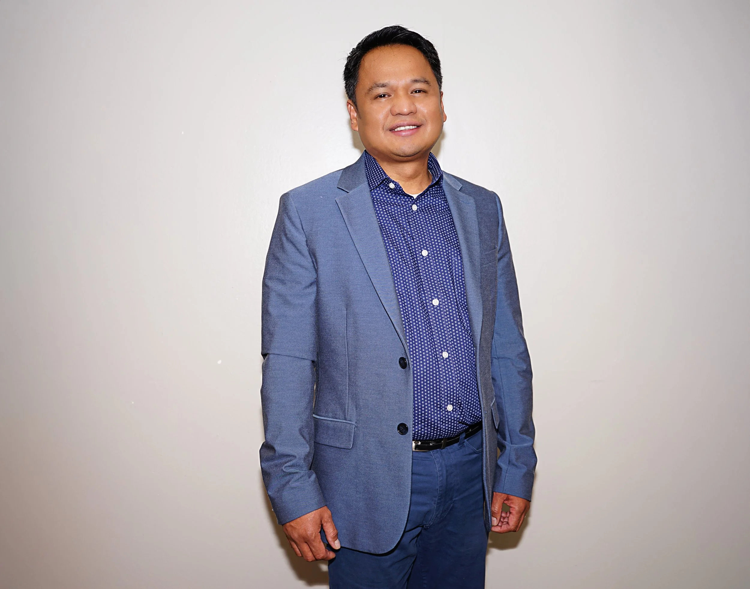 A man in a blue suit standing against a plain white wall, smiling at the camera.