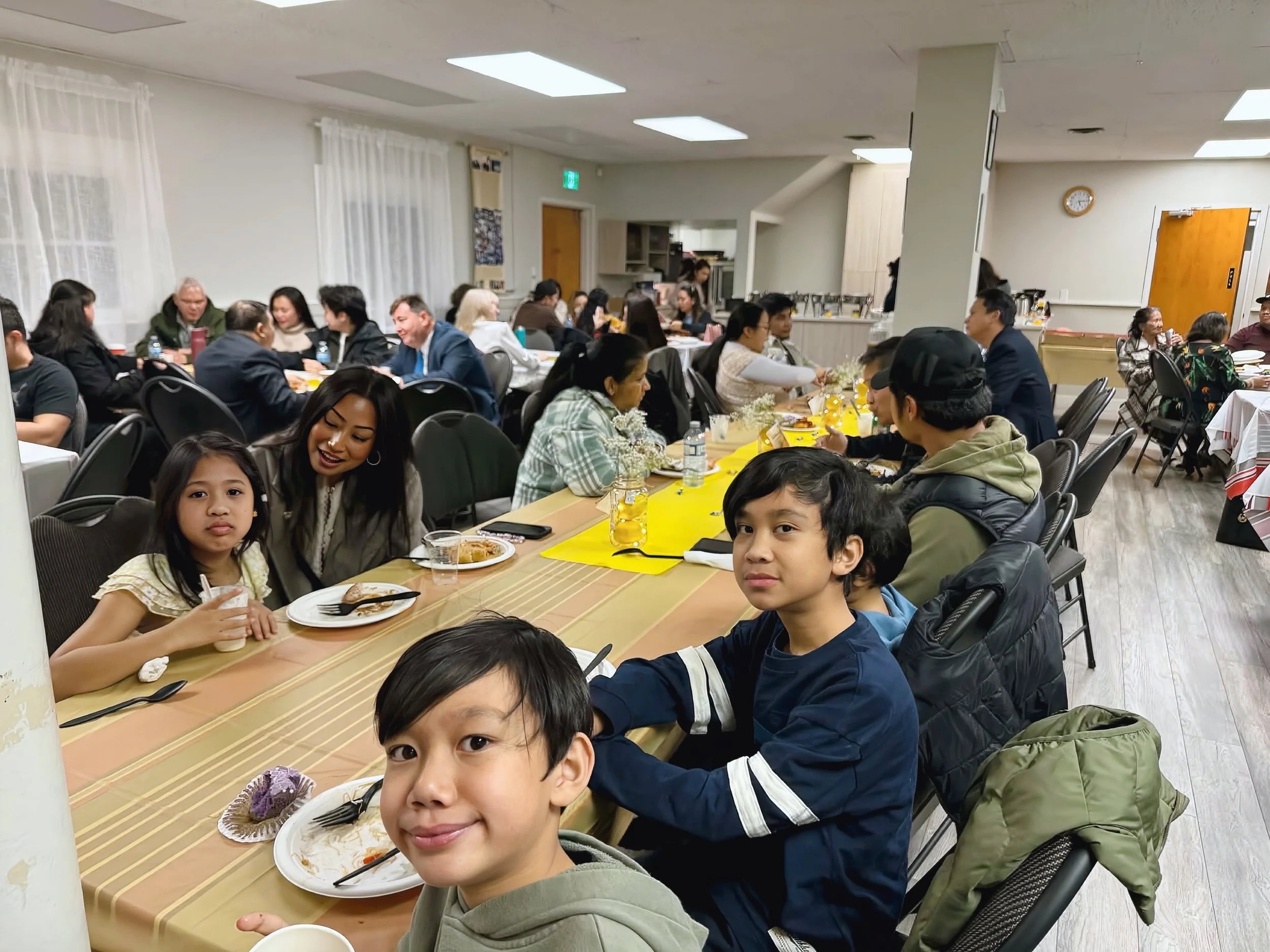 A group of children and adults seated at a banquet-style table in a large, well-lit room, enjoying a meal.