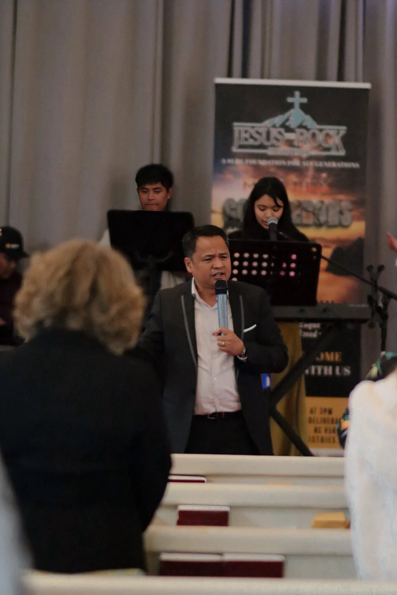 A man speaking into a microphone at a church or religious event, with a band or choir performing in the background.