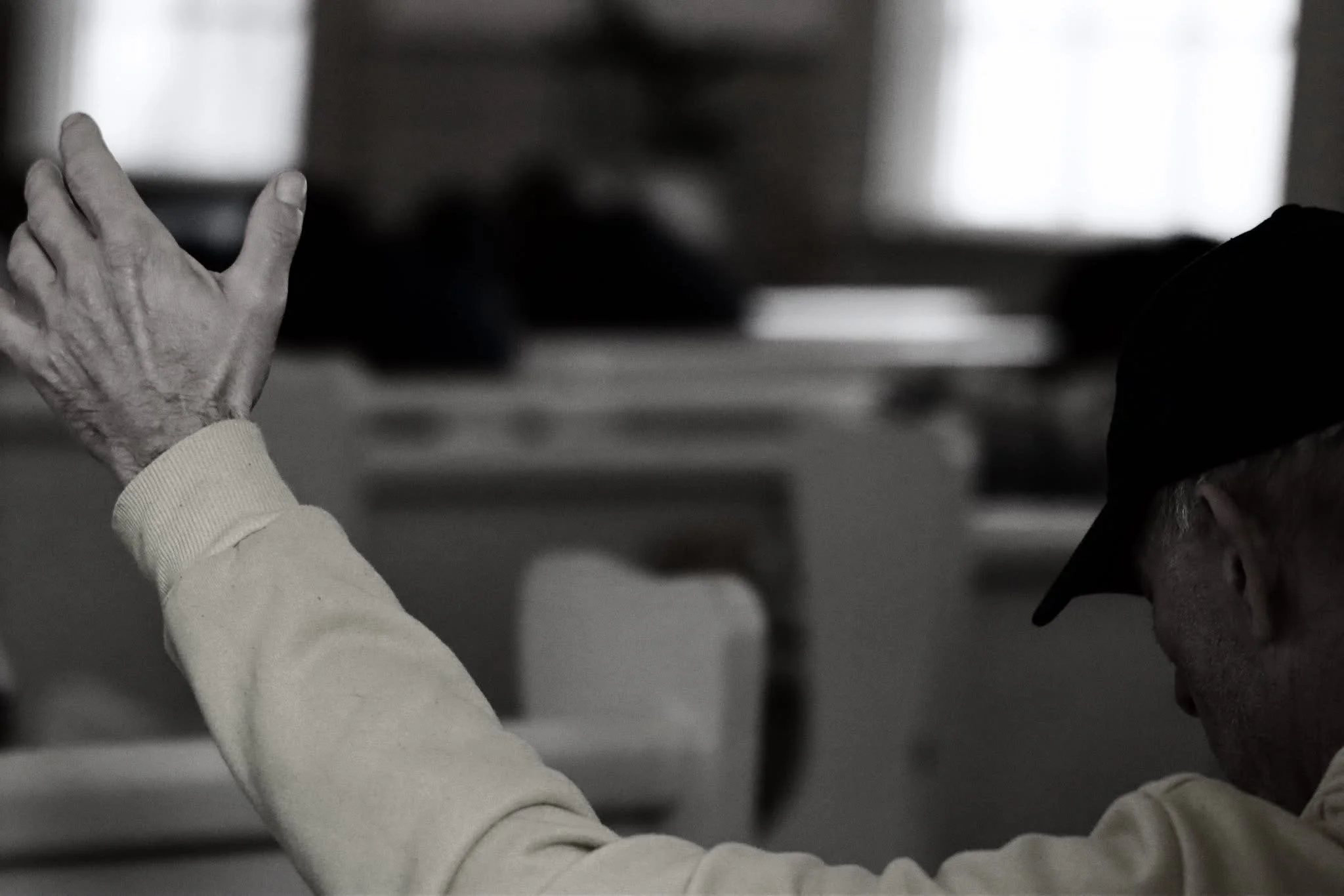 A senior man wearing a black cap and a beige sweatshirt raising his left hand in a gesture inside a dimly lit room.