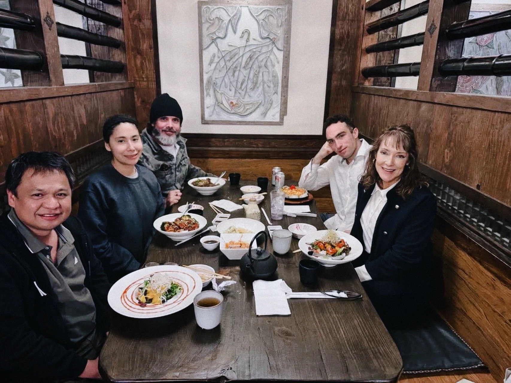 Six people sitting around a dining table in a restaurant, enjoying a meal with various dishes, including bowls of soup, salads, and a cake, with wooden walls and a sculpture in the background.