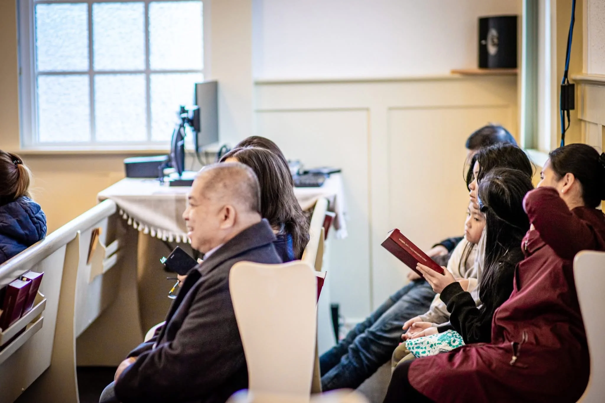 People sitting in a small church or classroom, some reading, some smiling, with a window and computer in the background.